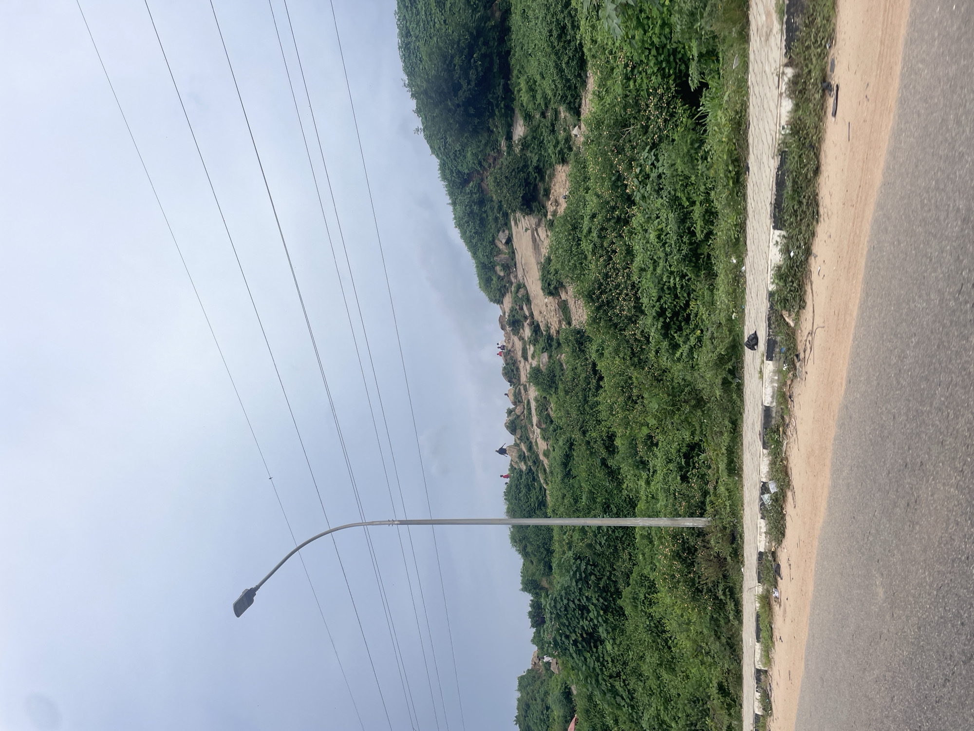 A street lamp stands tall on the left side of the image, its curved arm extending towards a cloudy sky. Several power lines stretch horizontally across the sky above the lamp and the landscape. In the background, a steep, rocky hill is covered in lush green vegetation. Several people are visible scattered across the hillside, some appearing to be climbing or standing on the rocks. A paved sidewalk runs horizontally across the foreground, bordered by a curb. The sidewalk is lined with patchy grass and scattered litter, including plastic bags and debris. A dirt shoulder separates the sidewalk from a paved road, which occupies the bottom portion of the image. The scene suggests an outdoor environment in a hilly, possibly semi-urban area.