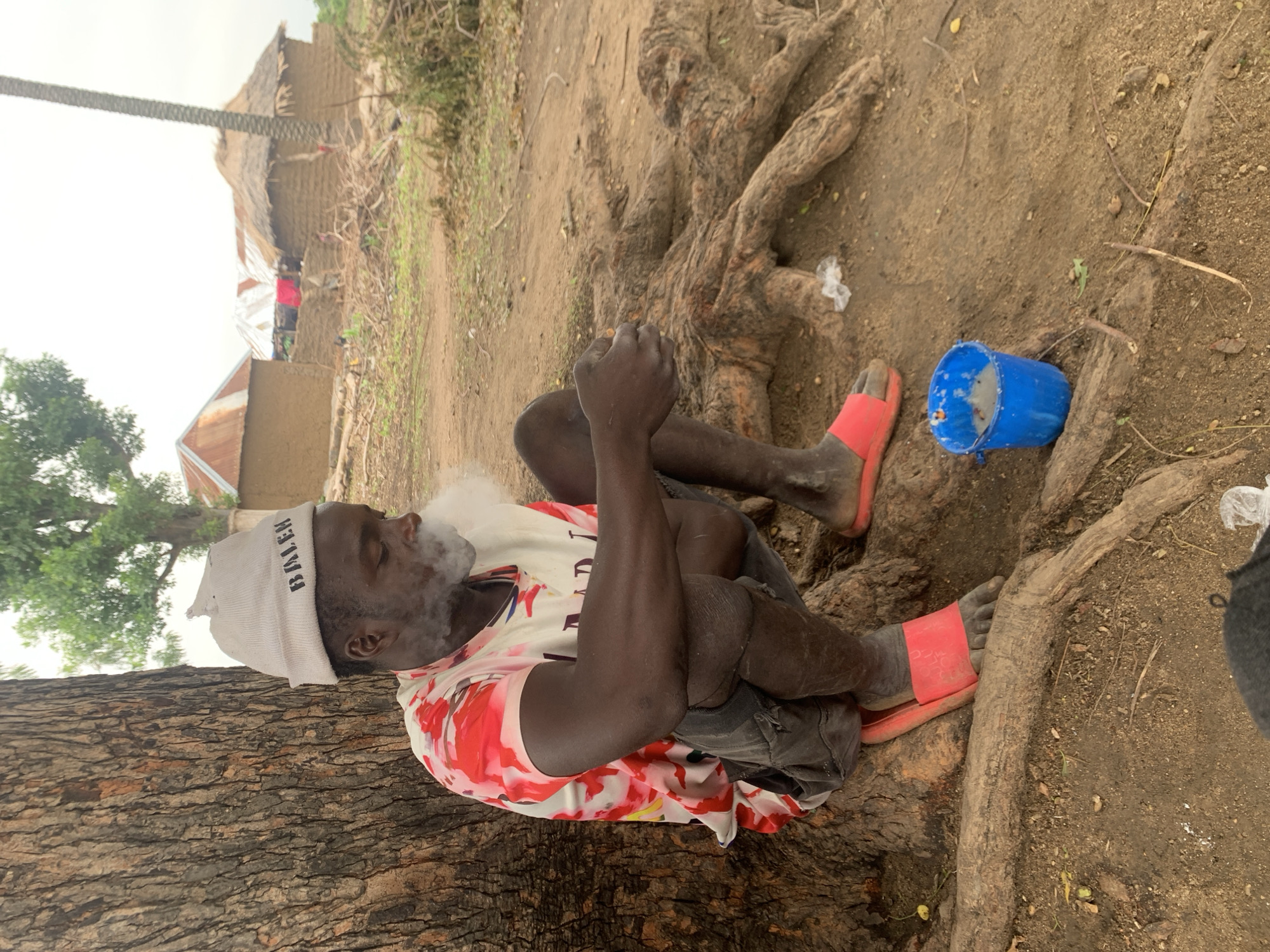 A Black man, wearing a grey knitted beanie with "BALEN" text, a red and white tie-dye t-shirt, and dark shorts, is crouched by the base of a large tree. He is smoking and exhaling a cloud of white smoke. His right hand is near his mouth, and his left hand rests on his knee. He is wearing red slide sandals. Beside him on the ground is a small blue bucket containing a light-colored substance, possibly food or liquid. The ground is covered in dirt and exposed tree roots. In the background, there are several mud-brick structures with corrugated metal and thatched roofs, indicative of a rural setting. The sky is overcast. This scene is located in Dull, Nigeria.