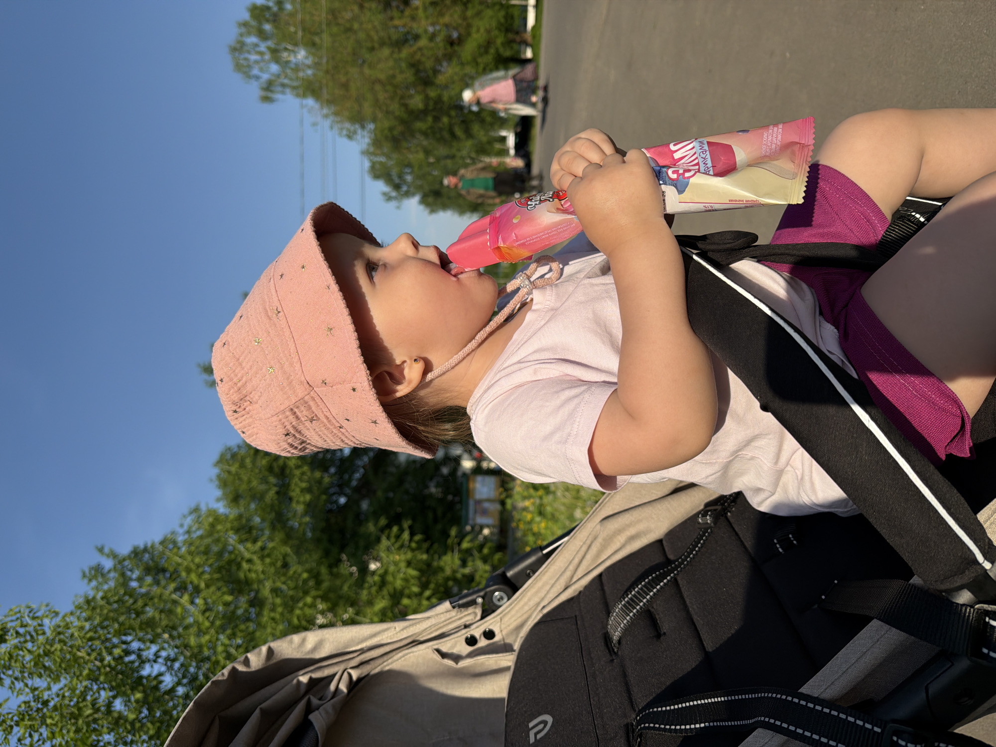 A young child wearing a pink hat and pink shirt is seated in a stroller. The child is consuming a packaged popsicle. The location is identifiable as Reshetnikovo, Russia, based on the Cyrillic writing on the popsicle packaging. The stroller has a black and beige color scheme with a visible 'P' logo. Green trees and a blue sky are visible in the background. Other people are distantly visible on a paved path.