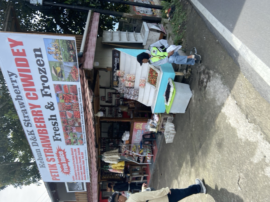 The image shows a street vendor stand in Rancabali, Indonesia (latitude: -7.122, longitude: 107.426). The stand is selling a variety of goods, including fresh strawberries, dried goods, and other snacks. It has a white sign above the roof, advertising the stand as "Kebun D&K Strawberry Petik Strawberry Ciwidey Fresh & Frozen Free Juice". The sign is red, white, and black, with a graphic of red strawberries on a white background. The vendor is selling his goods from behind a counter. There are two people in the image, one is a man wearing a yellow safety vest and blue jeans, sitting down by the counter and the other person is a man with a grey hooded jacket, dark blue pants and black shoes, walking away from the stand. There are two white plastic containers next to the counter, one of them containing a white plastic container filled with blackberries, and the other is empty. There is also a wooden table to the left of the counter with various products and two small wooden stools behind it. Behind the vendor, there is a large wooden structure that is possibly another stand, with a white sign that states "Kasih" with a small red sign to the left of it. On the left side of the structure, there are various goods hanging on hooks and another empty wooden table. The stand is located on the side of a paved road, with a white line separating it from the road. There is a green grassy area on the right side of the image.