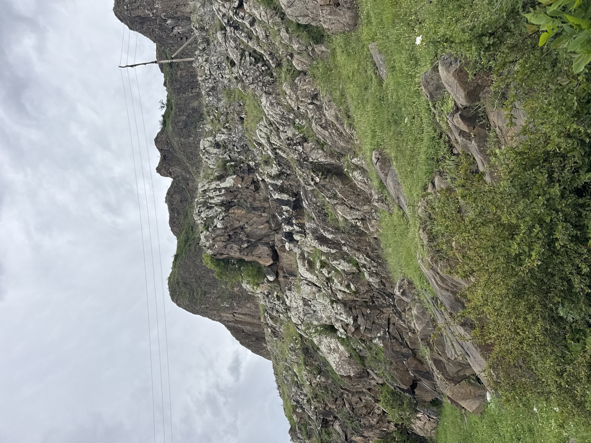 The image shows a rocky mountain landscape under an overcast sky. The mountain is covered with varying amounts of grass and sparse vegetation. A power line stretches across the sky. The image was taken in Каменномостское сельское поселение, Russia.