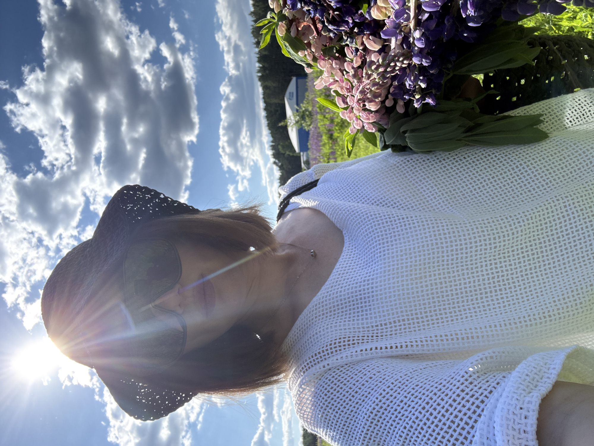 A female individual is depicted in a selfie-style photograph, standing in a field of blooming lupine flowers in Chaikovsky, Russia. The sun is positioned directly behind and slightly to her left, casting significant lens flare across the upper left quadrant of the image and providing strong backlighting to the subject. She wears a dark, wide-brimmed, woven hat and dark, oversized sunglasses. Her attire includes a white or off-white open-knit/crochet sweater with a wide neckline, and a thin chain necklace with a small dark pendant. Her hair is dark brown and appears to be bob-length. She holds a large, freshly-picked bouquet of multi-colored lupines, primarily in shades of pink, purple, and light blue, with prominent green foliage. The bouquet appears to be held in a dark, woven basket or bag, partially obscured. The background features more extensive lupine fields, a dense line of coniferous trees, and a distant light-colored building with a blue roof and blue horizontal detailing, under a blue sky with scattered cumulus clouds.