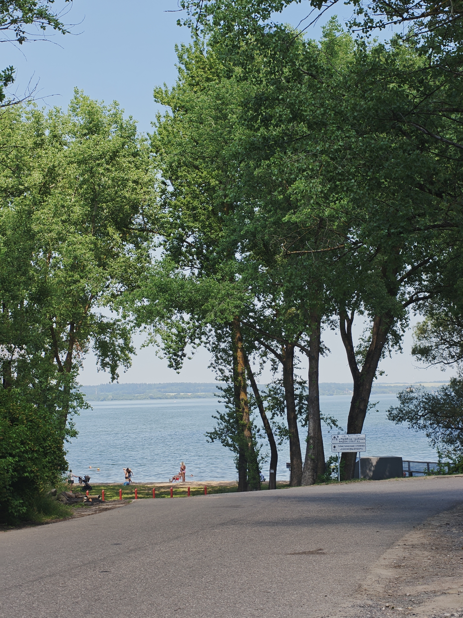 A paved road curves away from the viewer towards a body of water, likely a lake, fringed by a sandy beach and lush green trees. Several people are visible on the beach, some wading in the water, others standing and interacting. A stroller is parked near the edge of the road. A signpost with text in Cyrillic script stands near the water's edge, indicating directions to various points of interest, including "ТУРИСТИЧЕСКАЯ СТОЯНКА" (Tourist Parking) and "ЗОЛОТАЯ РЫБКА" (Golden Fish). The sky is a clear blue, with the sun casting shadows on the road. The overall scene suggests a recreational area on a warm day.