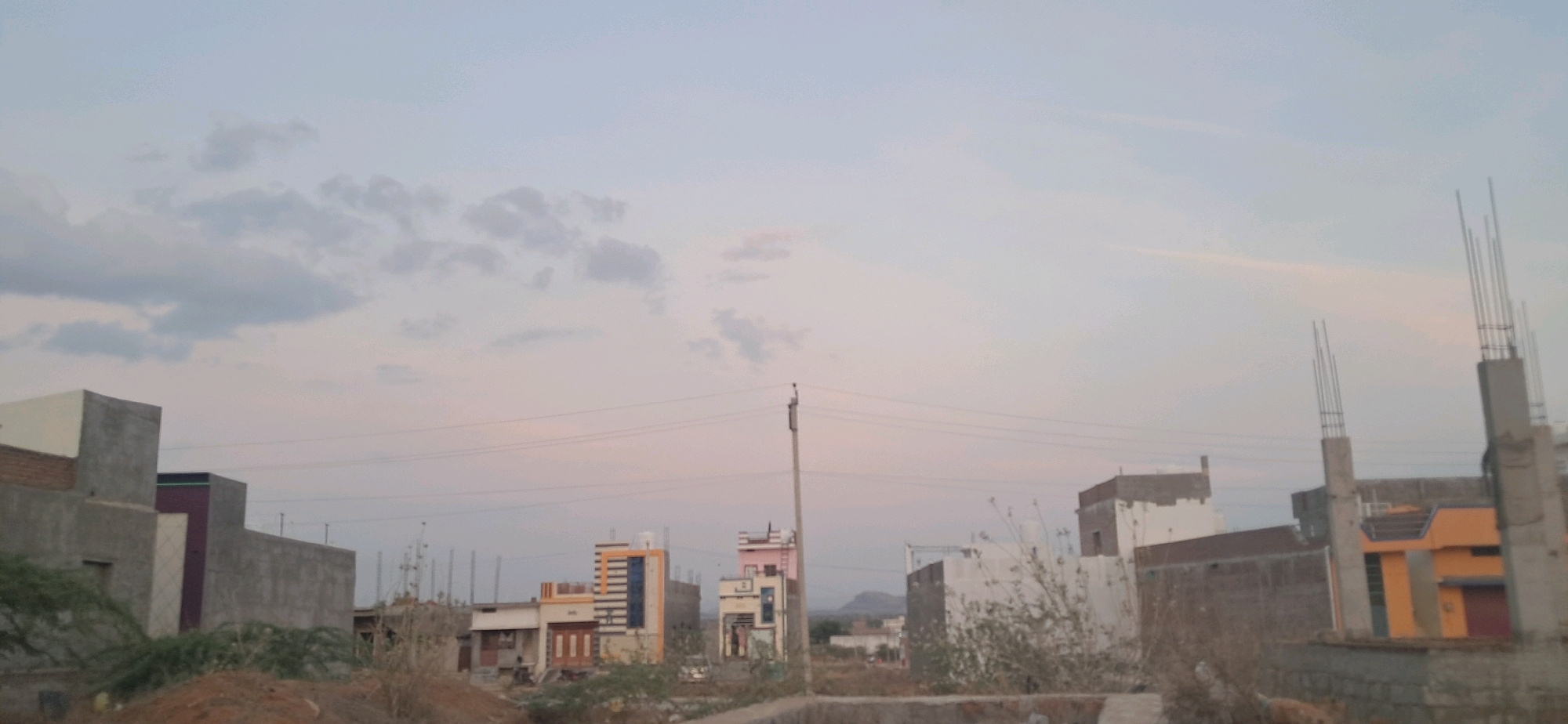This is a medium shot of a developing residential area in Rayachoty, India, captured during twilight. The sky is a pale, overcast blue with hints of pink near the horizon, and scattered, wispy clouds are visible. In the foreground, the ground is uneven, covered with dry grass, dirt, and some small shrubs.
The scene is dominated by a collection of houses and buildings in various stages of construction. On the left, a partially constructed building has exposed brickwork and a gray concrete facade. Next to it, a completed single-story house with a white-painted upper section and brick lower section is visible. Further into the mid-ground, several other houses are present. One striking building features a colorful facade with orange and blue accents, and striped windows. Another has a pink-painted upper story. To the right, several buildings are under construction, characterized by exposed concrete pillars with protruding rebar. One of these unfinished structures has a bright yellow and orange facade.
Power lines stretch across the frame, supported by utility poles. In the distance, a faint outline of hills or mountains can be seen against the horizon. There are no people visible in the image. The overall impression is one of ongoing development and residential growth in a semi-arid environment.