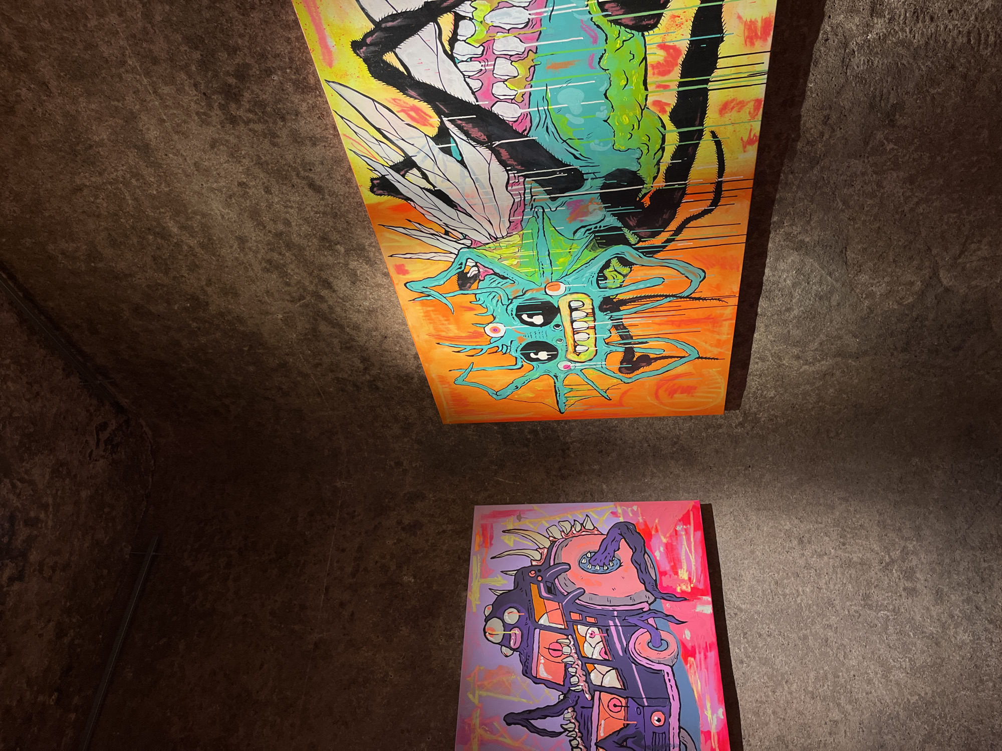 The image shows two pieces of contemporary art hanging on a dark, uneven stone wall. The upper piece is an orange-toned painting depicting an upside-down, stylized monster with green and blue accents, black appendages, and a white, wing-like feature. The lower piece is a pink and purple-toned painting of a machine-like creature. The wall appears to be part of a cave-like space. Based on the distinctive stone walls and the style of art, the location is possibly in Lisbon, Portugal, in a gallery or other art space. No people or interactions are visible in the image.
