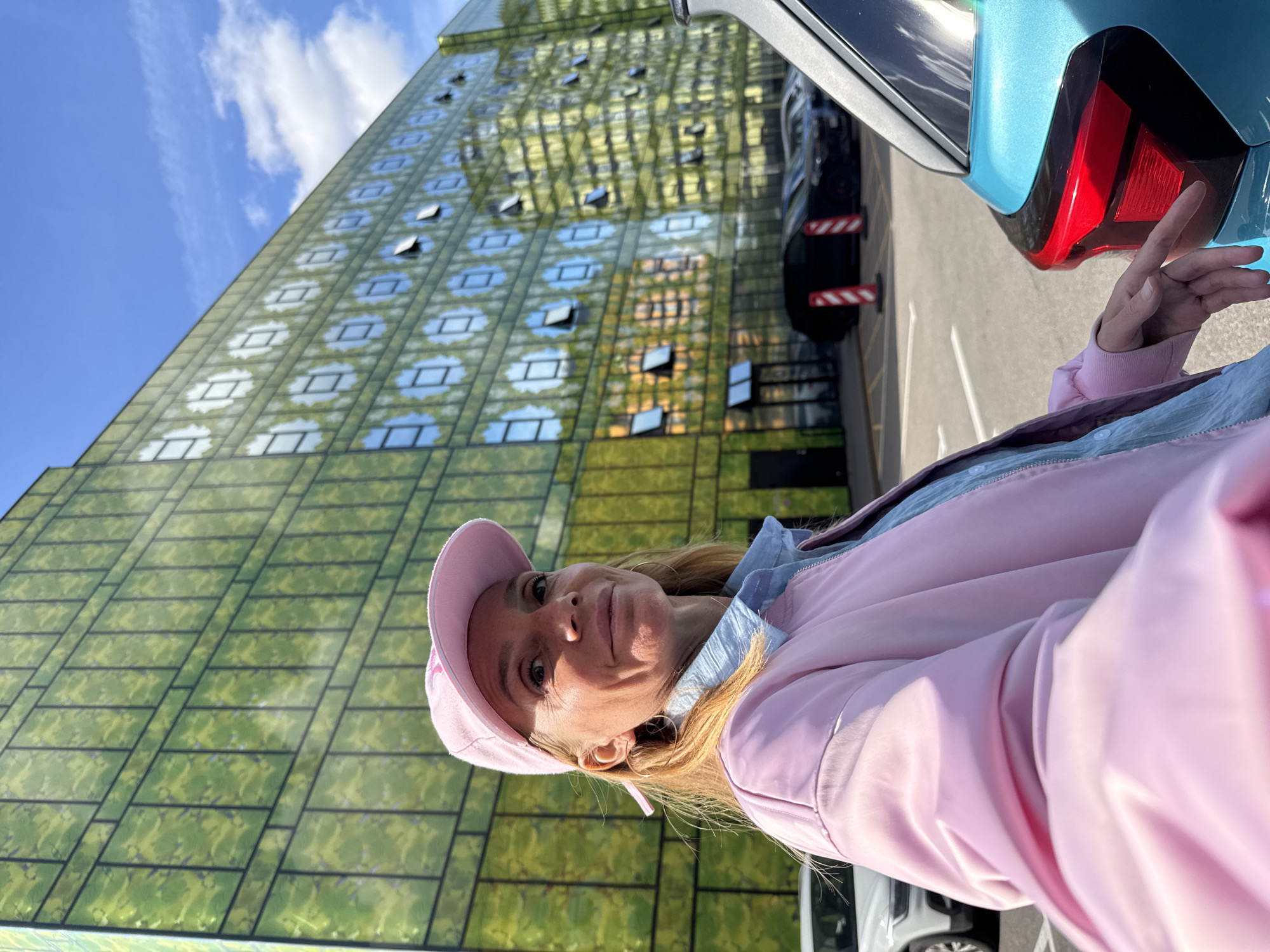 In this portrait-oriented, outdoor photograph, a woman with blonde hair is seen from the chest up, wearing a pink baseball cap and a pink jacket over a light blue collared shirt. She is looking towards the camera with a slight smile. She has hoop earrings and light makeup. The background features a modern, glass-facade building with a greenish tint, reflecting the sky and surrounding environment. Some windows on the building are open. To the right of the woman, the rear of a teal-colored car is visible, specifically its taillight and bumper. The scene appears to be a parking lot or outdoor area next to the building. The lighting suggests it is daytime, with the sun casting shadows and highlighting the reflective surfaces. The sky above is blue with scattered white clouds. The overall impression is one of a casual, possibly sunny day outing. The location is identified as Saint Petersburg, Russia. There is no visible text in the image.