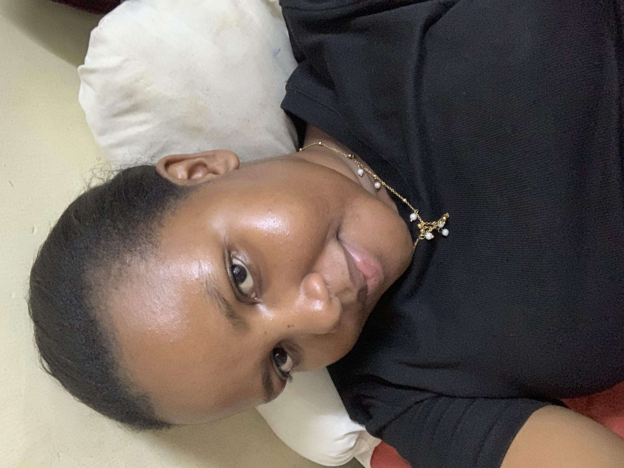 A medium shot depicts a woman lying on a white pillow against a beige-toned surface. She is wearing a black top and a gold necklace with small white bead-like adornments. The woman gazes at the camera. The background is undetailed. The location in Dar es-Salaam, Tanzania, cannot be determined from the given image.