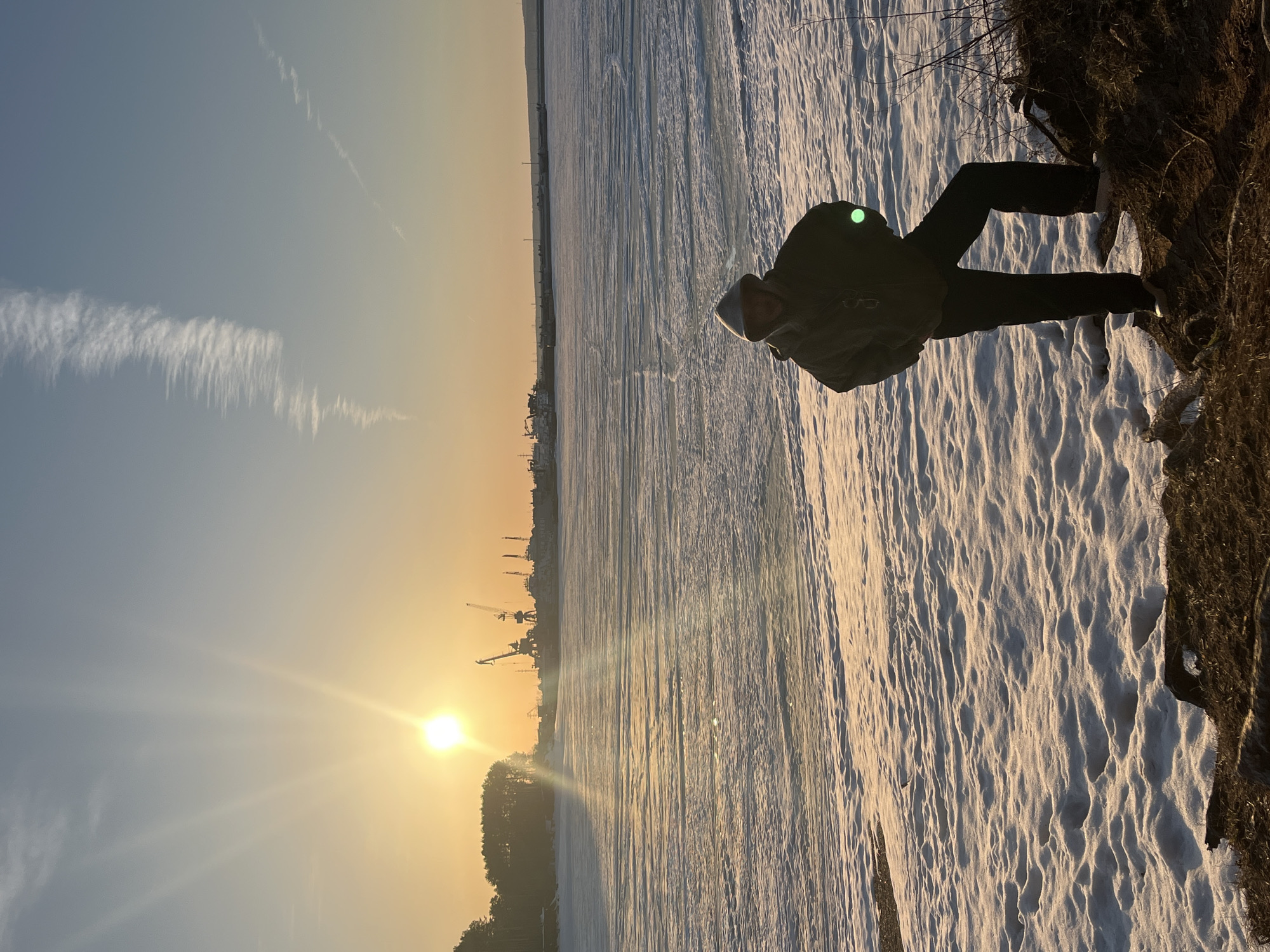 The image shows a person standing on a snow-covered ground near a bank of land with vegetation. The person is wearing dark pants and a jacket with a hood. In the background, a sunset illuminates a snow-covered field or body of water. Silhouetted against the sunset are trees and structures that appear to be cranes and industrial buildings, indicating a city skyline. The picture was taken in the city of Chaikovsky, Russia.