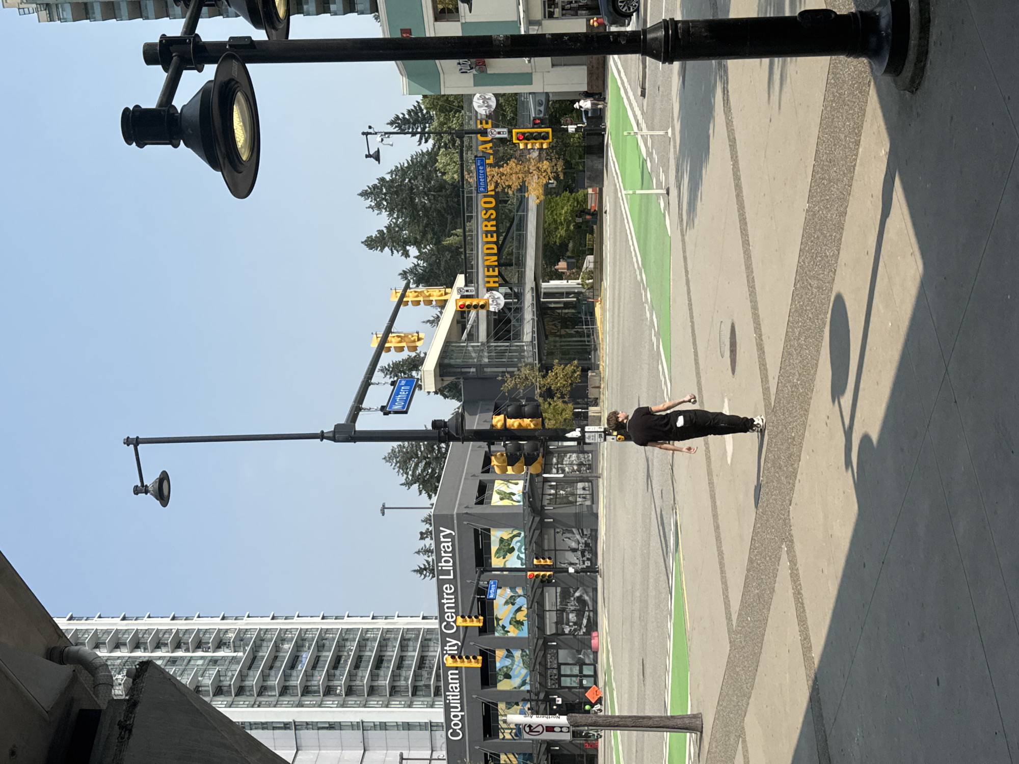 The image depicts an urban street scene in Coquitlam, Canada, specifically at the intersection of Northern Avenue and Henderson Place. In the foreground, a person wearing a black t-shirt and dark pants walks away from the camera across a paved pedestrian area with a patterned inlay. To the left of the pedestrian, a grey concrete structure, likely an elevated walkway or overpass, casts a shadow.
The background features a modern, grey building with the words "Coquitlam City Centre Library" prominently displayed in white lettering. The library has large windows and exterior murals depicting stylized green figures. Across the street, a pedestrian overpass spans the road, with a sign that reads "HENDERSON PLACE" in large yellow letters, and smaller signs indicating "Pinetree Place." Traffic lights are visible at the intersection.
Further in the background, a tall, modern apartment building with numerous balconies stands to the left, and a line of mature evergreen trees forms a backdrop to the right of the overpass. The sky is clear and blue, indicating a sunny day. Street lamps and traffic signals are integrated into the urban infrastructure. The street itself is marked with green lanes, likely designating bicycle paths.