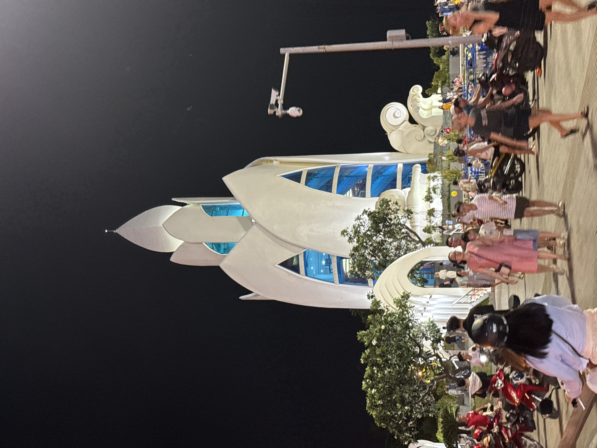 The image shows a night scene in Nha Trang Ward, Vietnam, with a prominent, modern building that appears to be a lotus flower design. The building has white, petal-like structures and bright blue windows. People are gathered in the foreground and around the building, some on motorcycles. The time of day is clearly night, indicated by the dark sky and artificial lighting illuminating the scene. Some people are dressed casually, and the overall atmosphere seems to be one of a public gathering or tourist attraction. The presence of motorcycles suggests a common mode of transportation in the area. The focus is on the unique architecture of the building and the activity of the people present.