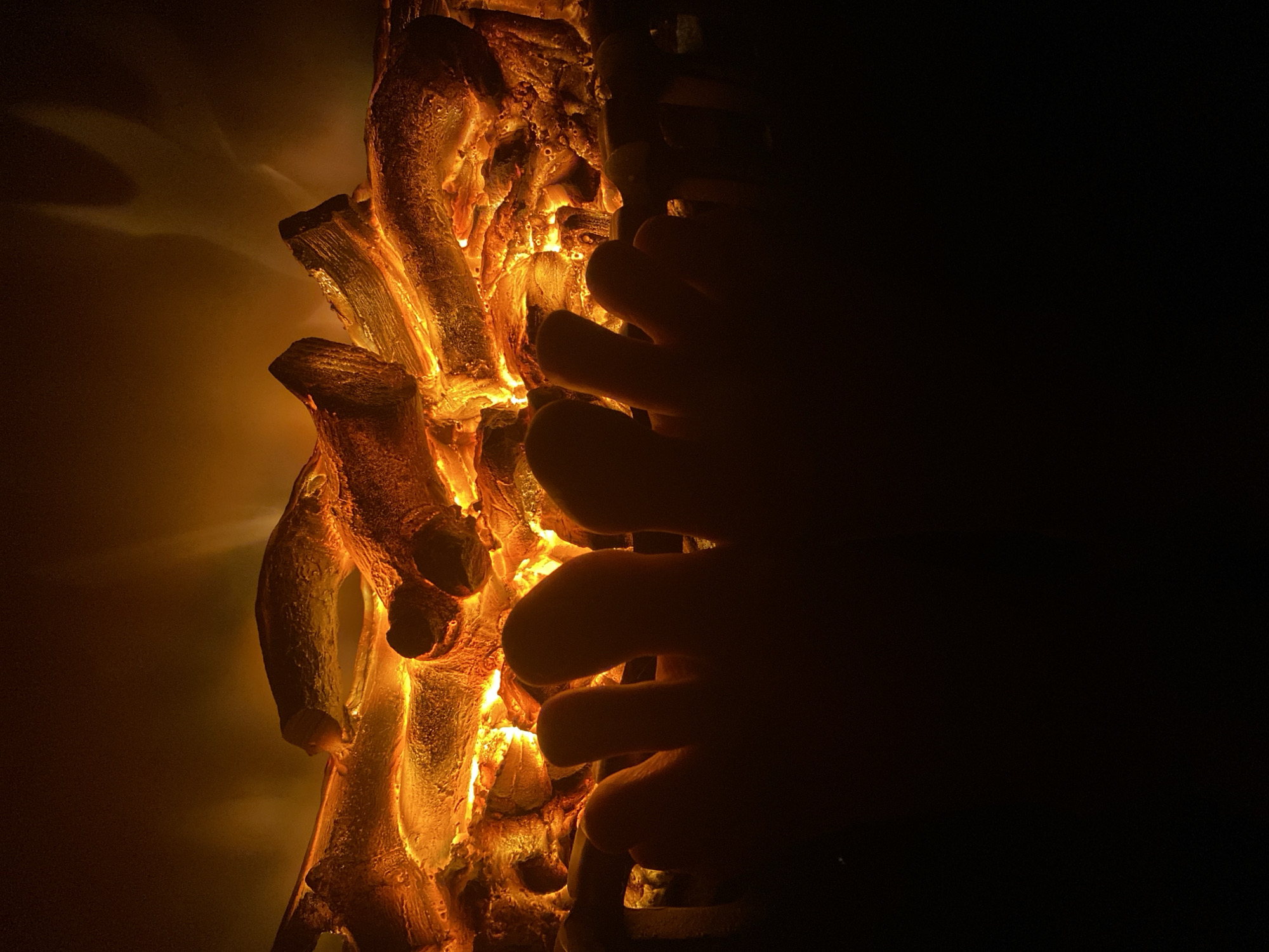 The image shows a close-up of a decorative object with a fiery, orange glow, possibly made of resin or a similar material to resemble charred wood or coal. The object is illuminated from within, creating a dramatic contrast between light and shadow. The texture of the material is rough and uneven, with visible details that suggest burning or intense heat. To the right of the object, a hand is partially visible in silhouette, reaching towards the illuminated structure. The background is dark, with the glow from the object casting light and shadows. The location of the image is not specified, and it is not possible to verify if it is in Novokuznetsk, Russia.