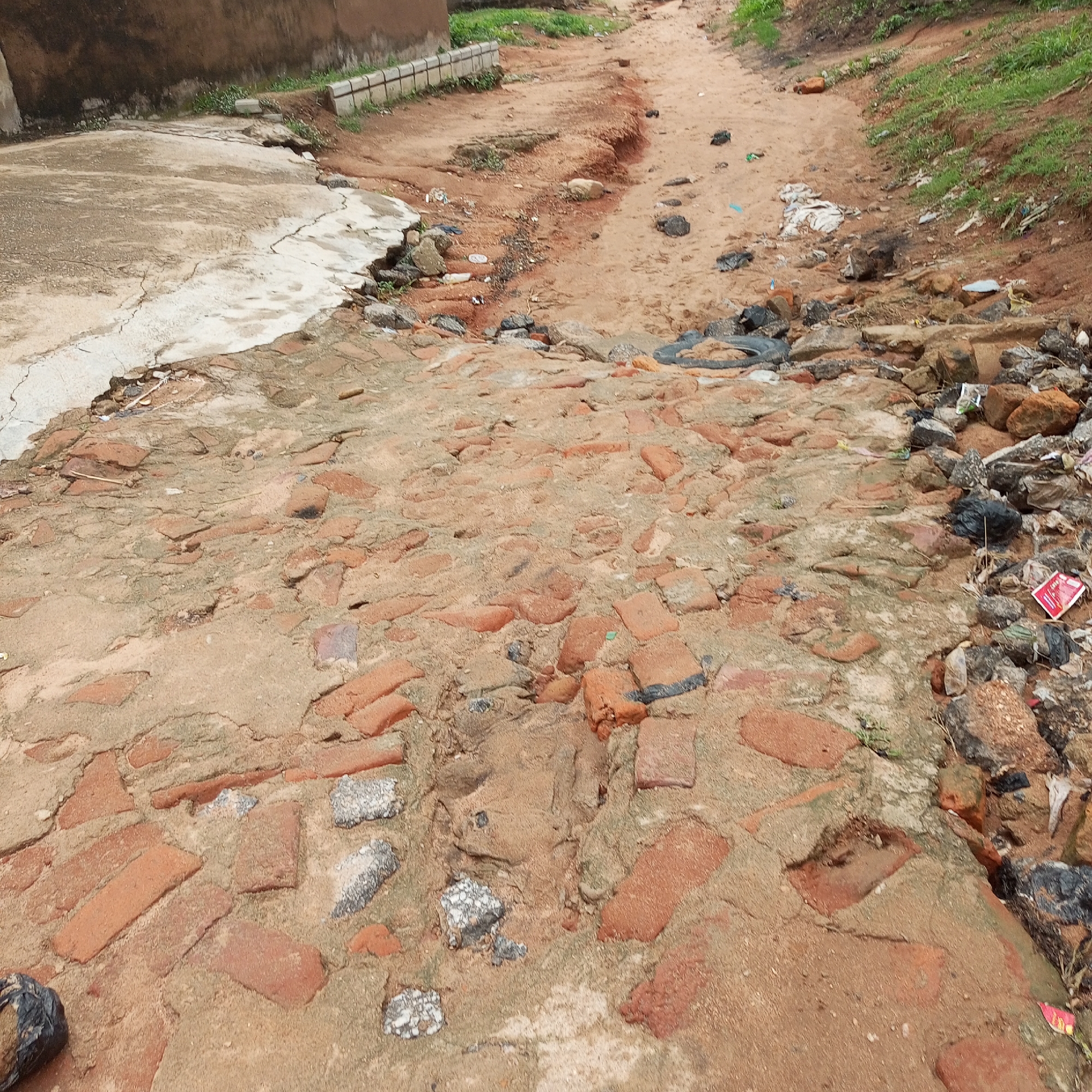 The image depicts a damaged dirt road with sections of brick paving. The road is uneven, with loose soil and scattered debris, including rocks, plastic bags, and what appears to be a discarded tire. The brickwork is partially intact and partially broken, with some bricks missing or dislodged, embedded in the soil. On the left side of the image, there is a section of cracked concrete pavement that slopes downwards towards the brick-paved area. In the background, a weathered building with a brown facade is visible, with a low concrete barrier along its base. Green grass grows along the sides of the road and embankment. The scene appears to be in a state of disrepair, characteristic of a poorly maintained urban or semi-urban environment. This scene is consistent with locations in Jalingo, Nigeria, where infrastructure can be subject to wear and tear due to weather and usage.