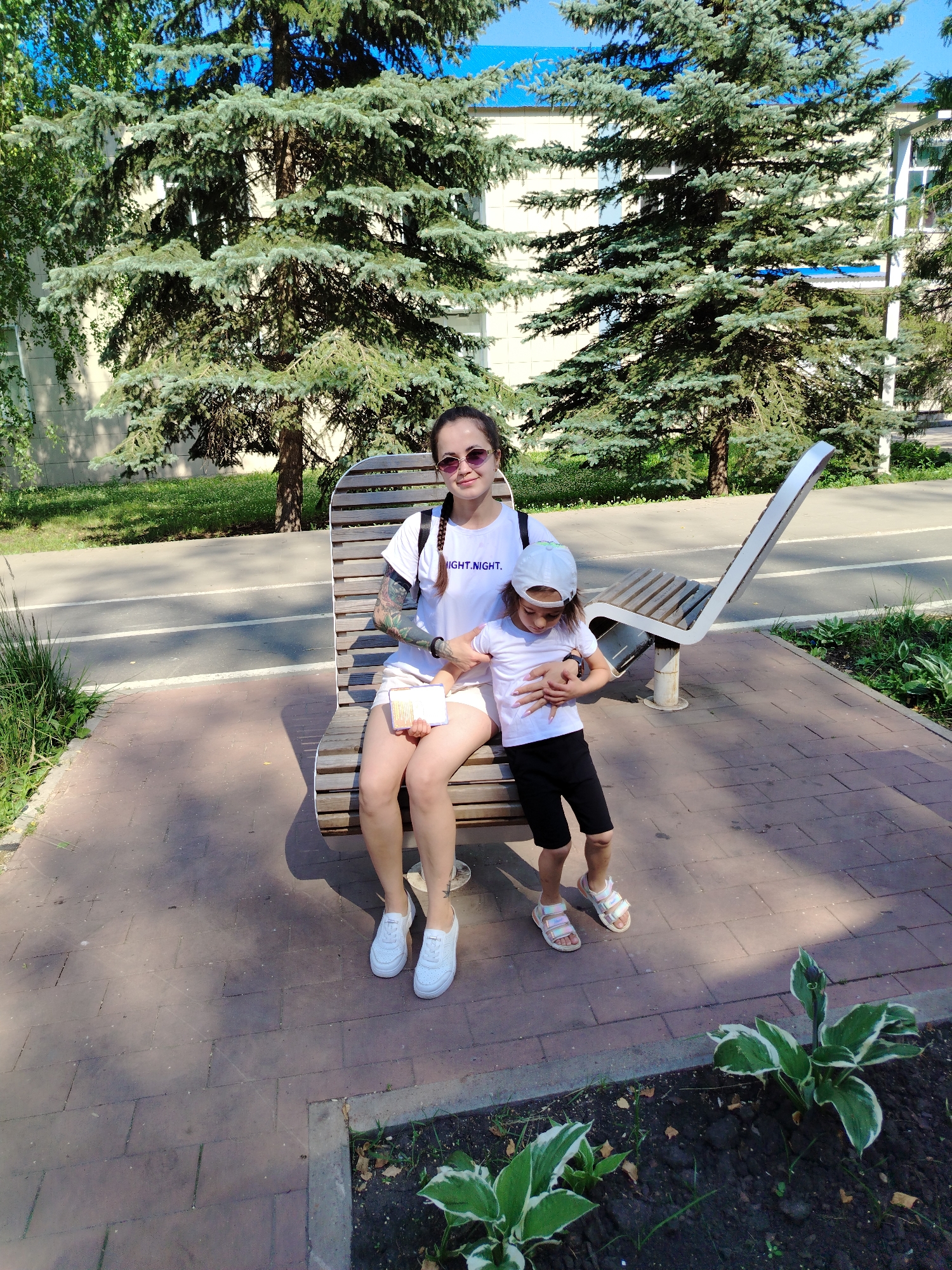 In Yanaul, Russia, a woman and a child are seated on a park bench under the shade of evergreen trees. The woman, wearing sunglasses and a white t-shirt with "NIGHT.NIGHT." printed on it, holds a small notebook in her lap. The child, dressed in a white t-shirt and black shorts, wears a white cap and rests their head on the woman's arm. The bench is made of wood and metal and is situated on a paved walkway adjacent to a grassy area and a bike path. The background features a light-colored building with blue trim and mature trees.
