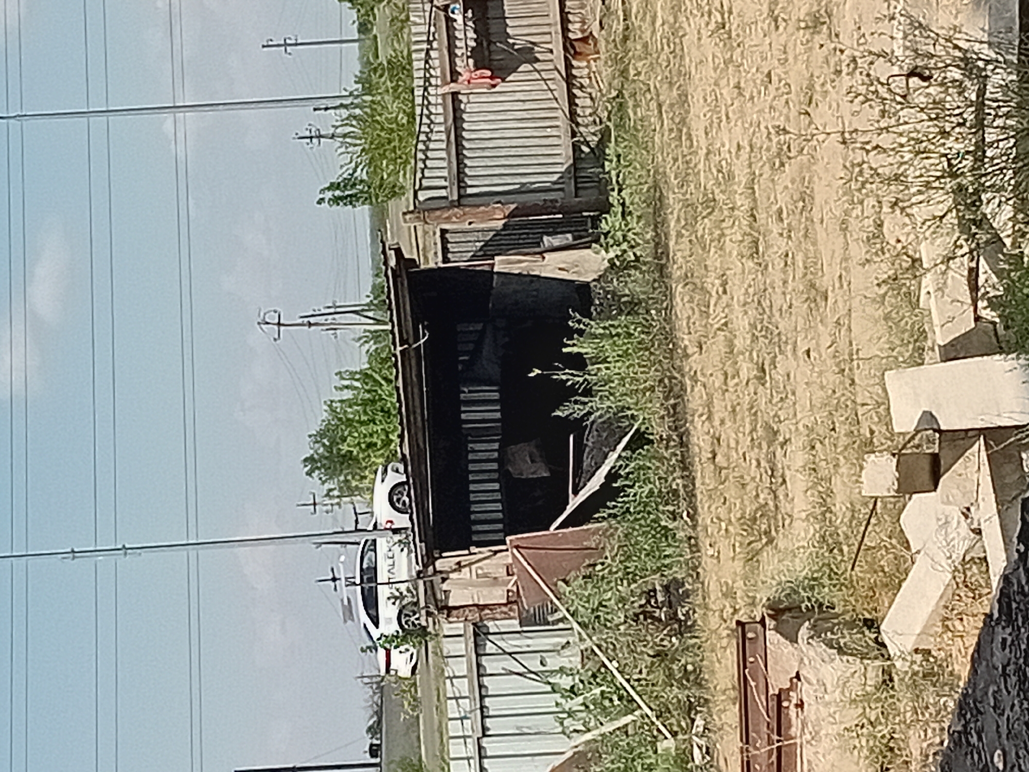In Semey, Kazakhstan, a white sedan with the logo "STALEKS" on its side is parked near a dilapidated metal shed. The shed appears to be constructed from corrugated metal and has a partially collapsed roof. It is surrounded by overgrown weeds and debris, including what looks like discarded metal framing and concrete blocks. A corrugated metal fence runs along the right side of the shed, with some red fabric hanging from it. In the background, utility poles with power lines are visible against a bright, hazy sky. The overall scene suggests an industrial or semi-abandoned area.