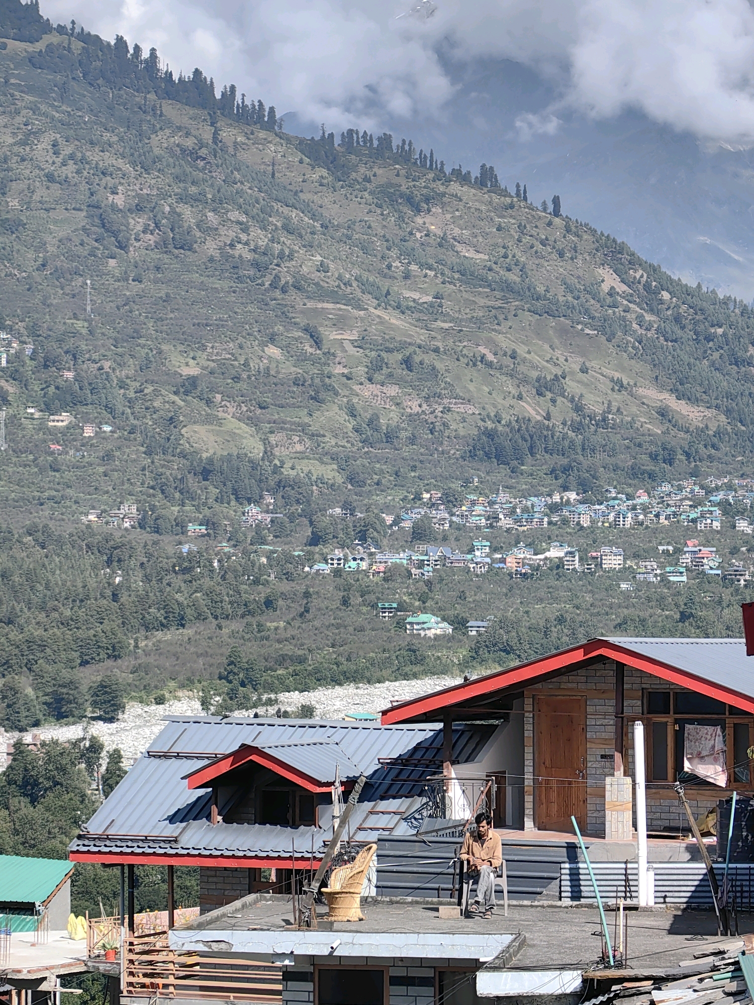 The image captures a scenic view of Vashisht, India, with a person sitting on a chair on a rooftop in the foreground. Behind the rooftop, a mountain covered with trees and houses is visible. The sky is partly cloudy.
In the foreground, a man wearing a brown shirt and grey pants is seated on a black chair on a concrete rooftop. The rooftop has a grey tiled roof with red trim, and a wooden door is visible on the building. Next to the man, there is a woven wicker chair.
The middle ground features a densely populated area with numerous buildings and houses scattered across the mountain slope. The houses are of various colors and architectural styles, with many featuring green or red roofs. Trees are interspersed throughout the housing area.
The background is dominated by a large, verdant mountain. The slopes are covered with a mixture of trees and smaller shrubs. Patches of bare earth and terraced areas are also visible on the mountainside. The upper part of the mountain is shrouded in clouds.
The overall scene depicts a residential area nestled within a mountainous landscape, typical of Vashisht, a popular tourist destination known for its hot springs and temples. The lighting suggests it is daytime, with the sun casting shadows.
