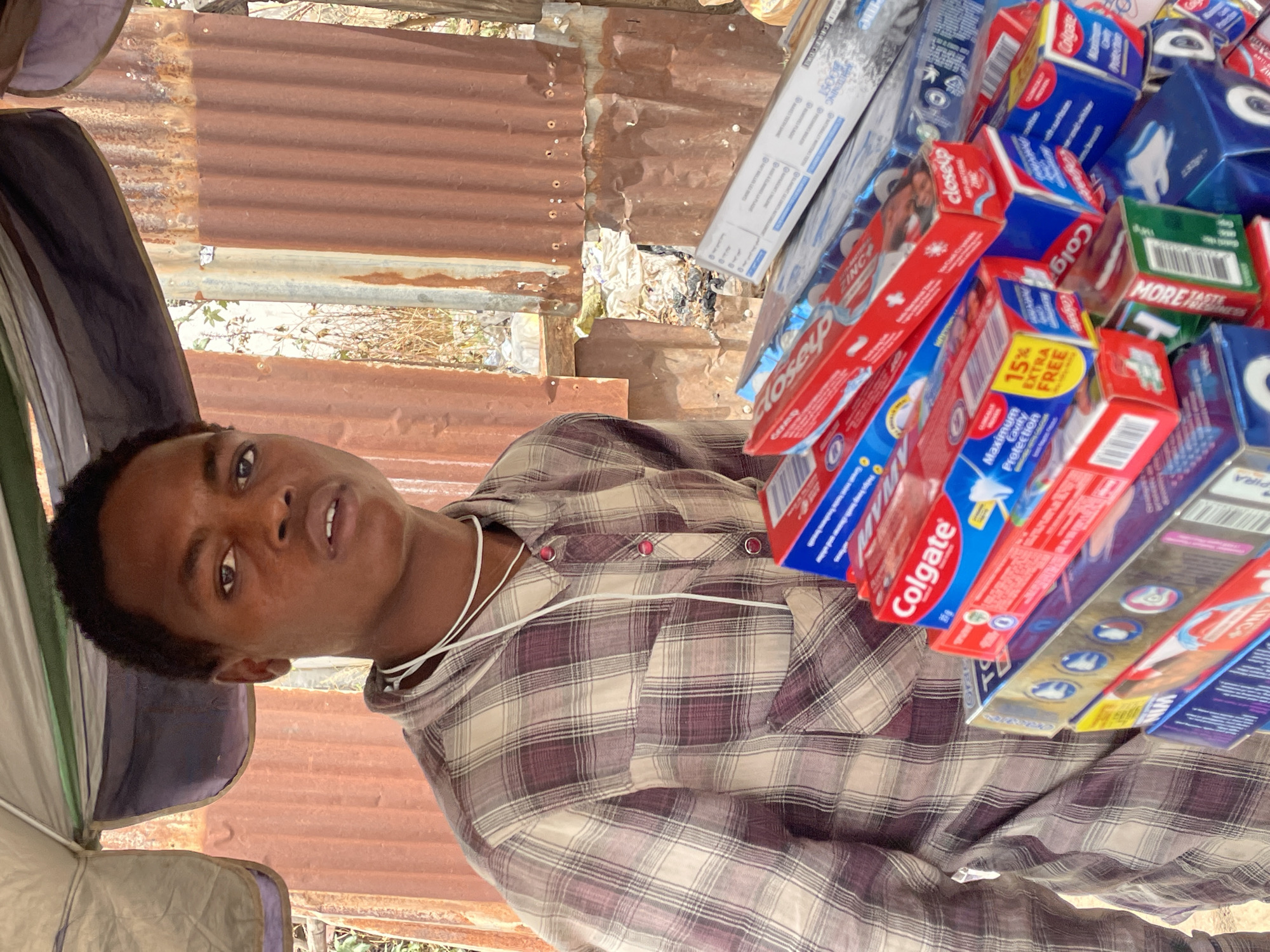 In Jos, Nigeria, a dark-skinned young man stands behind a pile of toothpaste boxes. He has short dark hair and wears a plaid shirt with a white string necklace. The boxes of toothpaste, including Colgate and Close-Up brands, are stacked haphazardly. Behind him is a corrugated metal structure and a glimpse of vegetation. He appears to be selling or displaying the toothpaste.