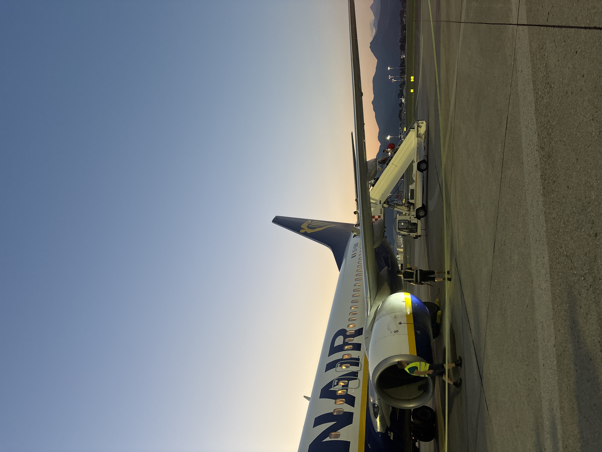 At Orio al Serio International Airport in Italy, during twilight, a Ryanair airplane is parked on the tarmac. A mobile stairs unit is attached to the aircraft, and several passengers are seen ascending. Two ground crew members, one wearing a high-visibility vest, are visible near the engine of the plane. The background shows airport lighting, other aircraft in the distance, and a mountain range silhouetted against the fading light of the sky. The scene is illuminated by the aircraft's landing lights and the ambient twilight.