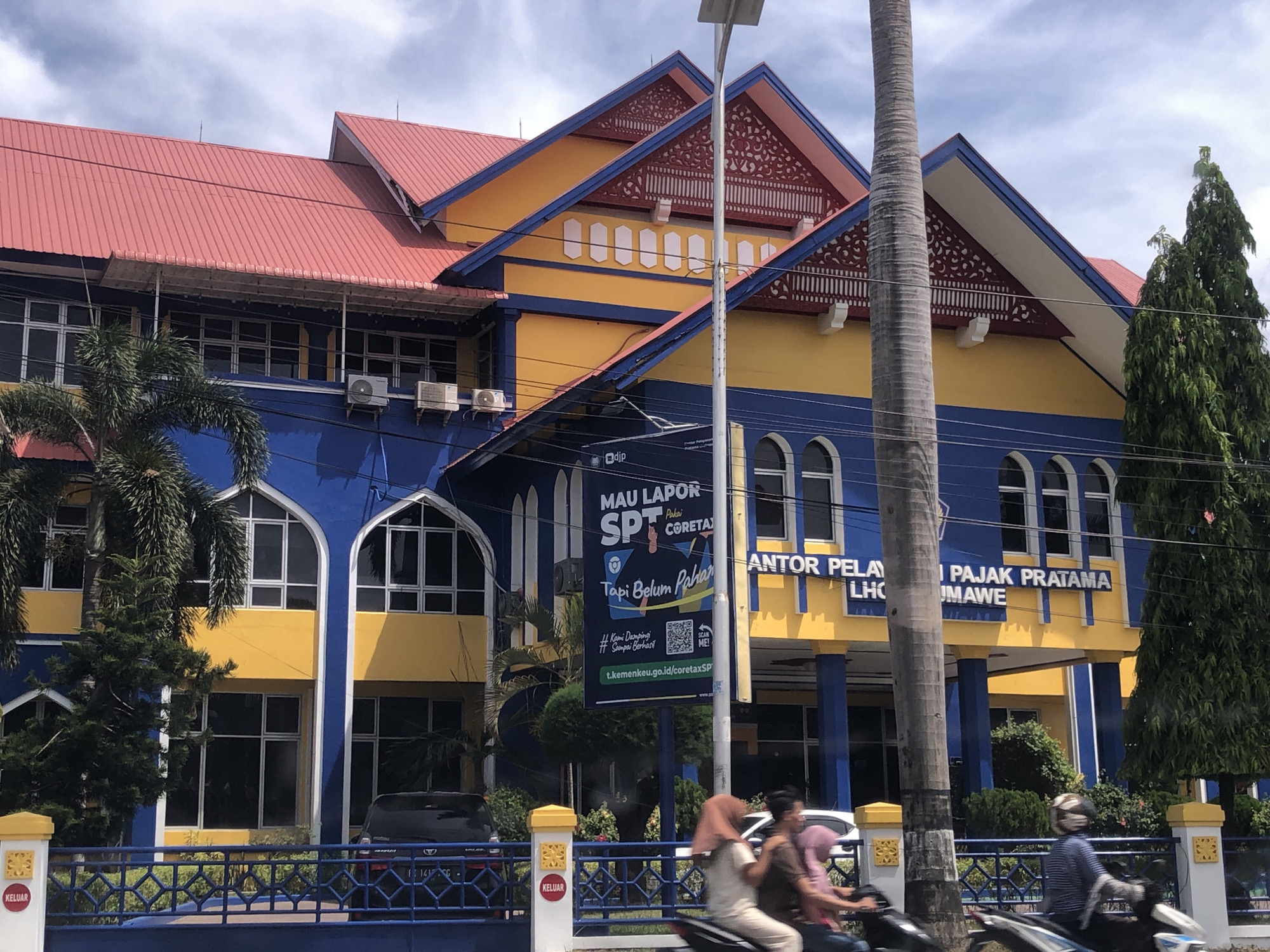 The image shows a colorful, three-story building in Lhokseumawe, Indonesia, which appears to be the local tax office. The building features a red roof, yellow and blue walls, and arched windows. A large banner in front of the building advertises tax services, featuring the words "MAU LAPOR SPT Pakai CORETAXI" ("WANT TO REPORT SPT Use CORETAXI"). The banner also includes a QR code and text "#Kami Dampingi Sampai Berhasil" ("#We Assist Until Success"). In the foreground, there's a blue fence, greenery, and a street with people on motorcycles passing by. A palm tree stands to the side of the building. The sky is overcast.