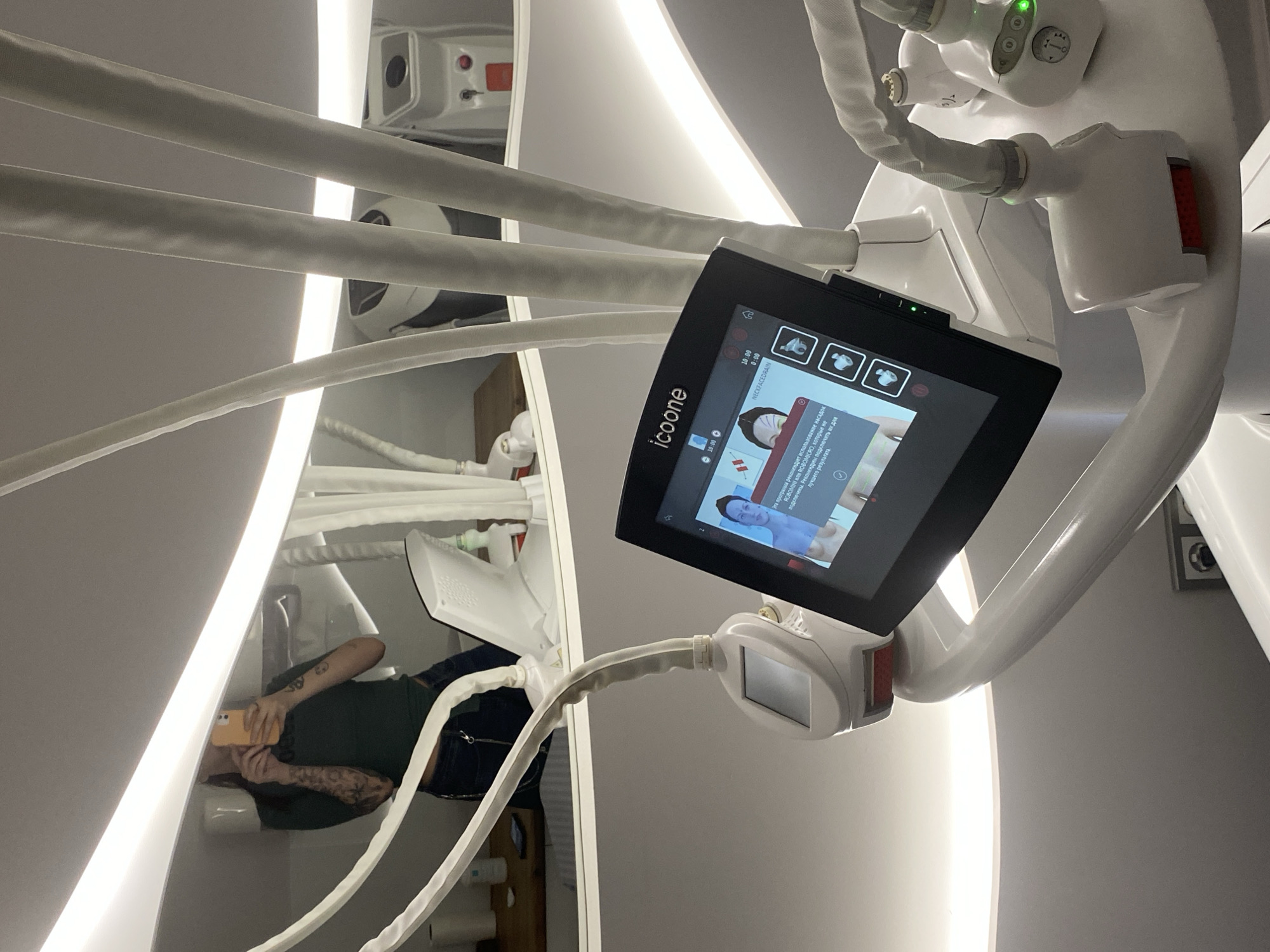 A low-angle shot shows an icoone medical device with a black touchscreen displaying text in Russian and icons, surrounded by white tubes and handles. The machine is located in a clinical room. Reflected in the glossy surface of the icoone device, a person in a green shirt and jeans is lying back, holding a yellow smartphone. The room has bright, built-in linear lighting, and other medical equipment is visible in the background. The city of Votkinsk, Russia, cannot be confirmed.