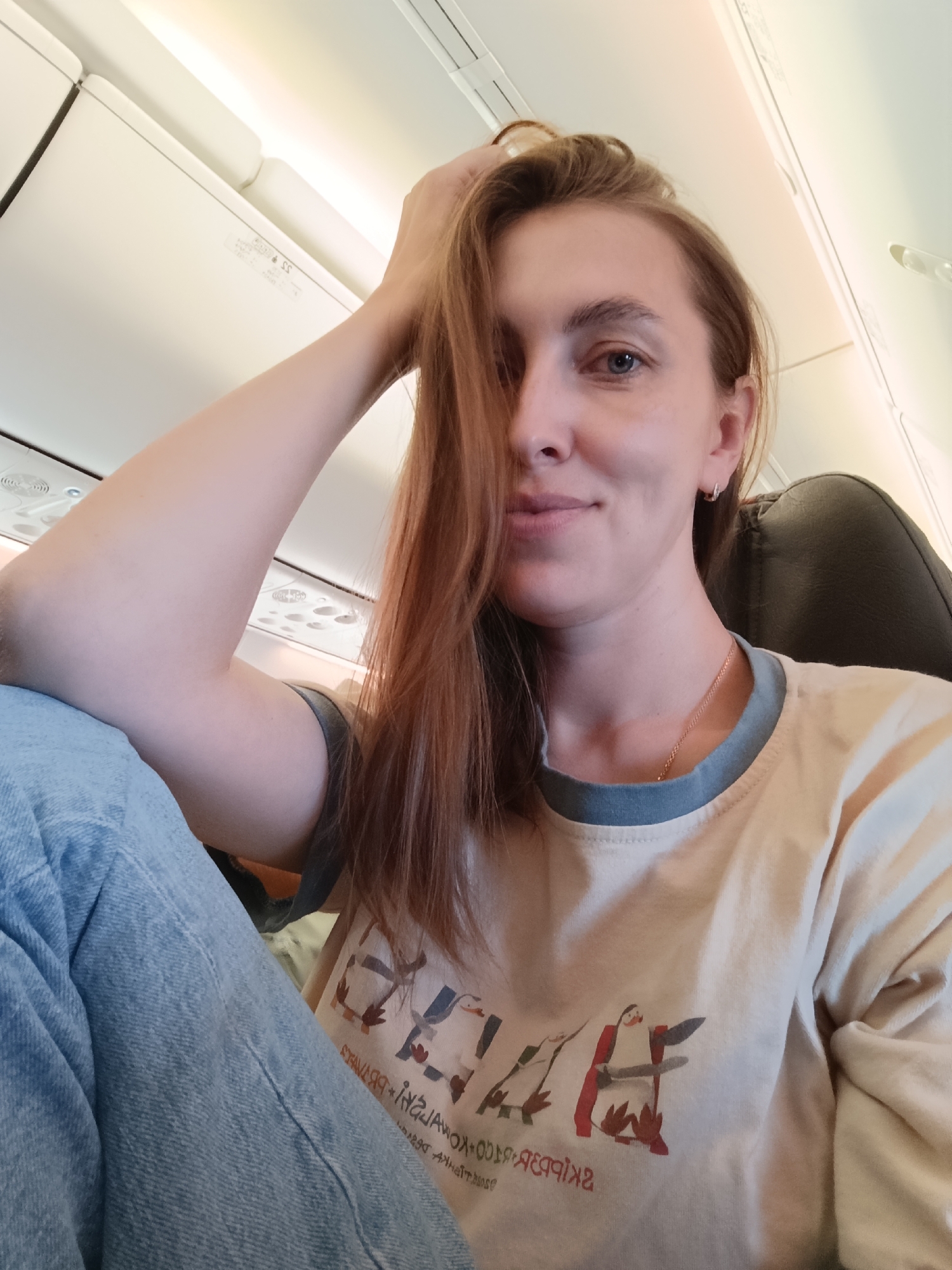 The image is a selfie taken from a low angle, showing a young woman with reddish-brown hair sitting on an airplane. She is wearing a light-colored t-shirt with a blue collar and blue jeans. Her right hand is resting on her head, partially covering her face. The background appears to be the interior of an airplane cabin, with overhead compartments and lighting visible. There are no discernible landmarks or elements that place the location specifically in Staroe Mart'yanovo, Russia.