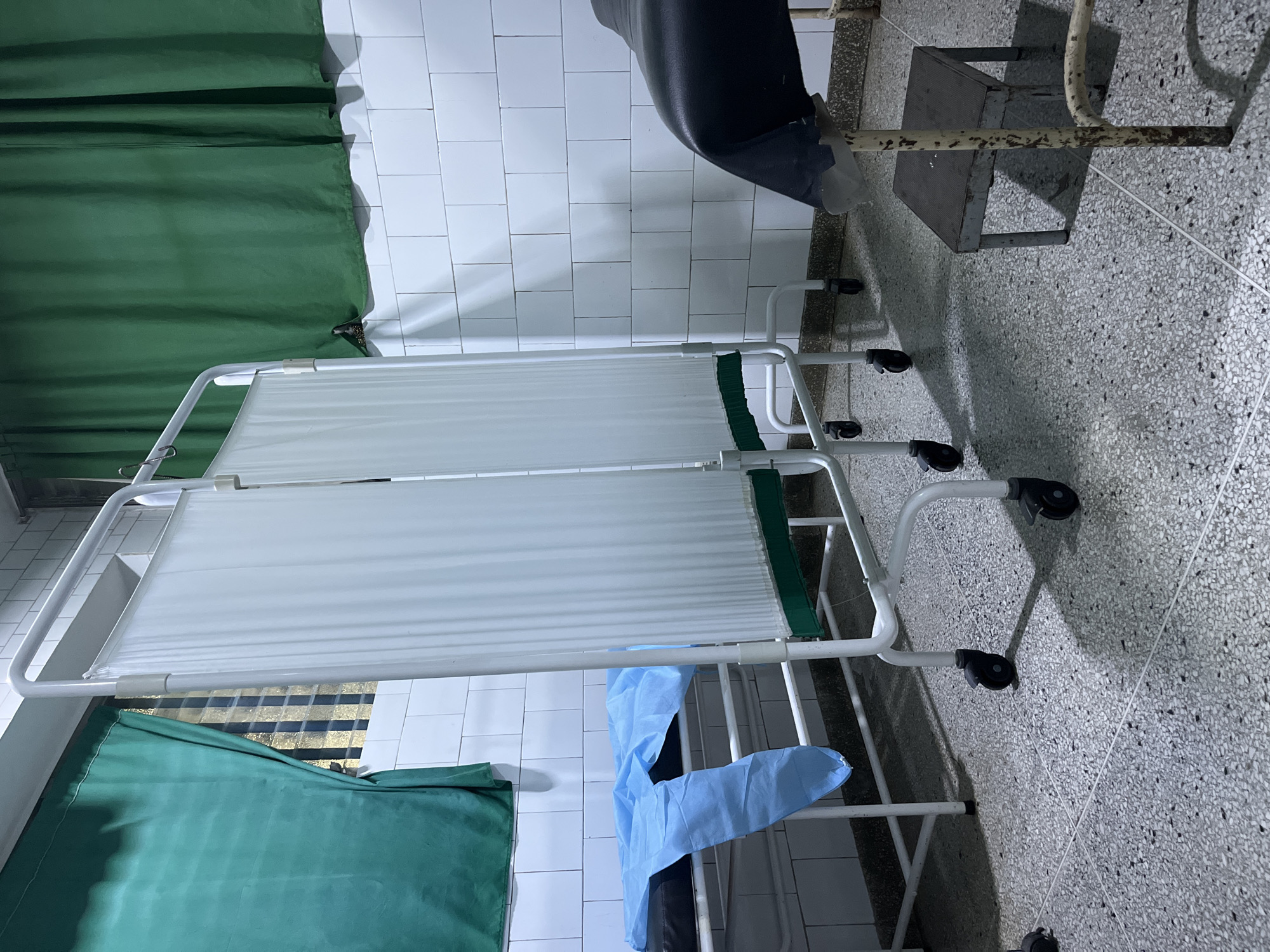 The image depicts what appears to be the corner of a room in a medical facility. The walls are tiled in white, and the floor is terrazzo. The scene is dominated by a mobile privacy screen with white fabric panels and a white metal frame with green accents. A green curtain is visible on the left side of the image. A discarded blue surgical gown or drape is partially draped over the lower part of the privacy screen. A dark chair and a small metal platform are in the upper right of the image. The specific location within Juan Griego, Venezuela, cannot be determined from the image. There are no people in the image.