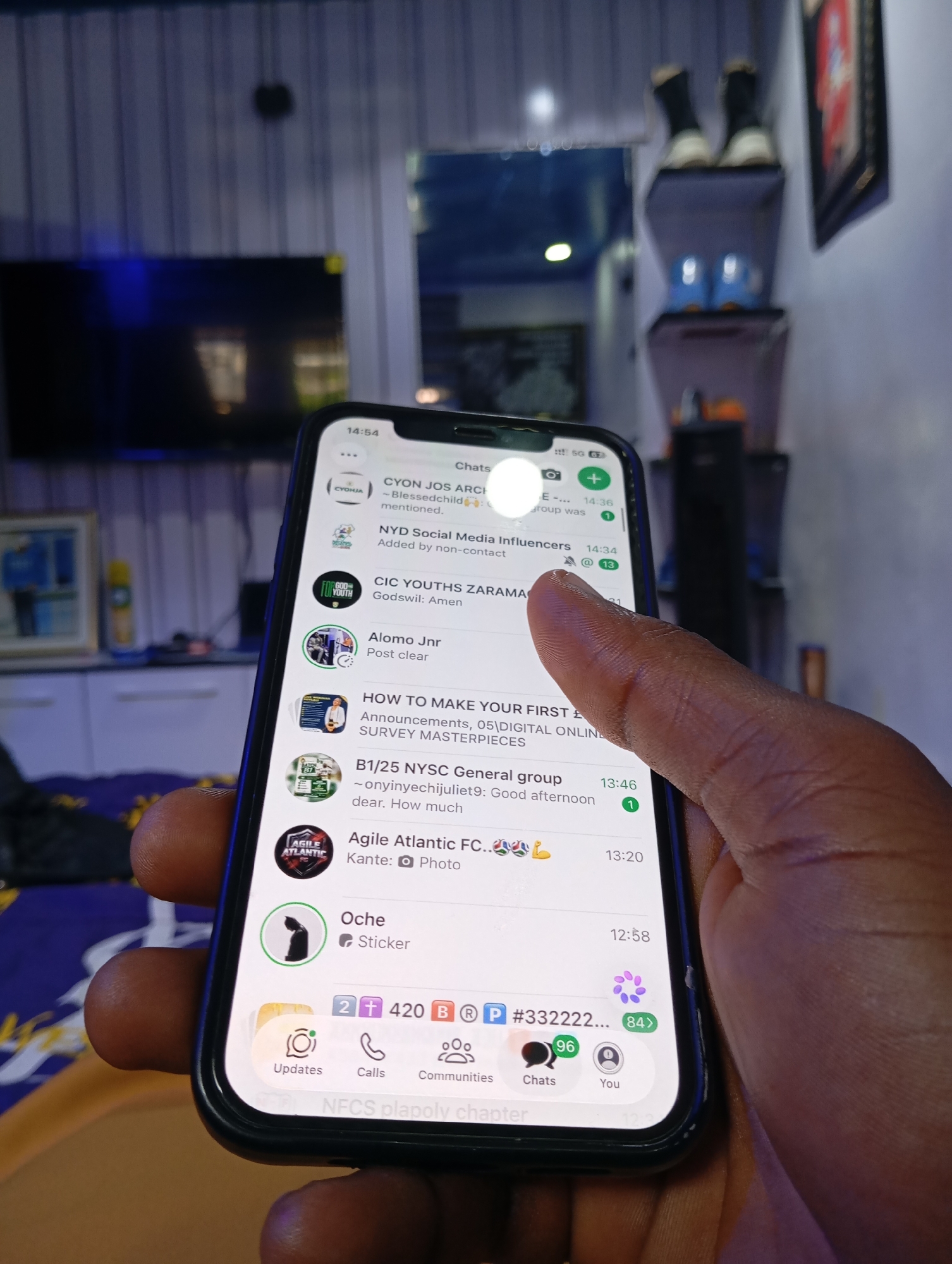 A person's hand is holding a smartphone displaying a messaging application, likely WhatsApp, in a dimly lit room. The phone screen shows a list of chat conversations, with the topmost visible chat belonging to "CYON JOS ARCHI". Other visible chat names include "NYD Social Media Influencers", "CIC YOUTHS ZARAMA", "Alomo Jnr", "HOW TO MAKE YOUR FIRST £", "B1/25 NYSC General group", and "Agile Atlantic FC.". At the bottom of the screen, navigation icons for "Updates", "Calls", "Communities", "Chats", and "You" are visible. The background of the room is blurred, showing furniture and what appears to be a television screen. The overall impression is of a casual, indoor setting.