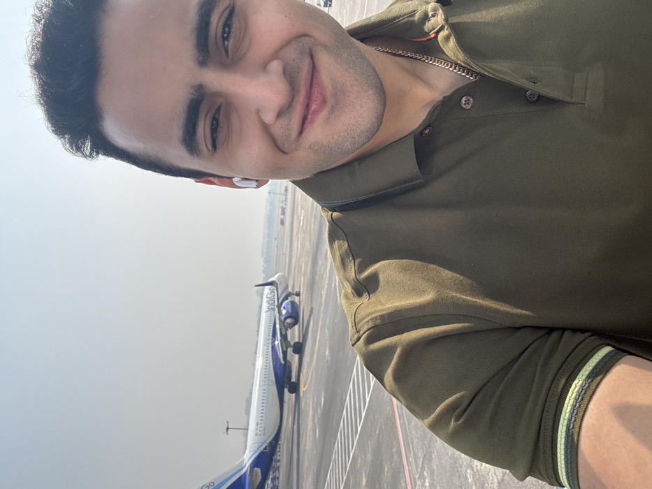 A man in an olive green polo shirt poses for a selfie on an airport tarmac. In the background, a blue and white Indigo airplane sits on the runway. The man has short, dark hair, fair skin, and a slight smile. He is wearing a necklace and an earpiece. The sky is hazy and the overall lighting suggests it's daytime. The picture is taken at an angle, with the man's face in the upper portion of the frame.
