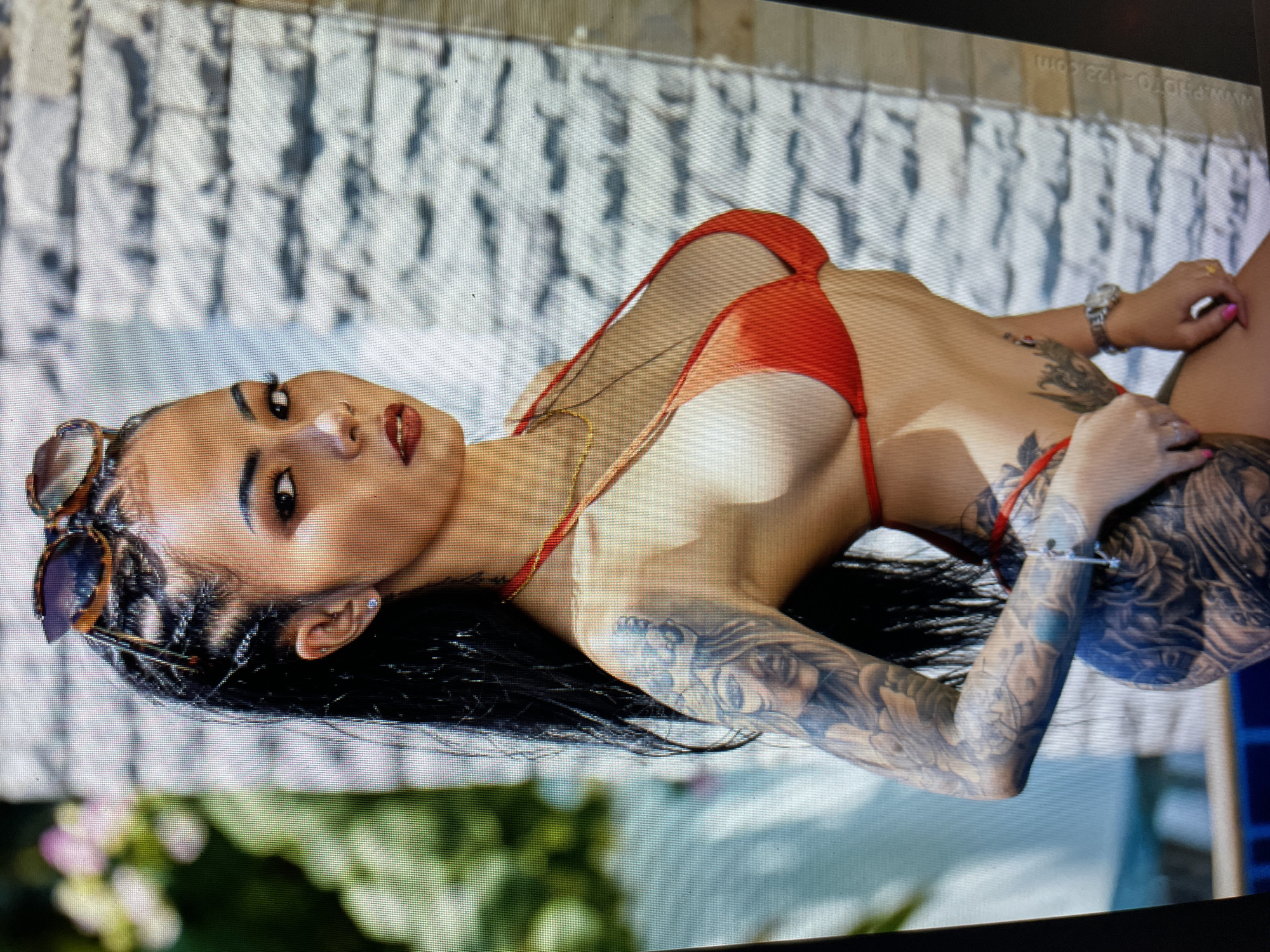 A woman is lying on her side wearing a red bikini. She has black hair styled in braids and long loose hair. She has sunglasses on her head. Her left arm is heavily tattooed with detailed designs. Her right arm and hand are partially visible, showing a wristwatch and gold ring. She has a gold necklace. She is lying on a surface next to a swimming pool, with a textured white wall in the background. The bottom left of the image shows blurry greenery. The location is Bacong, Philippines.
