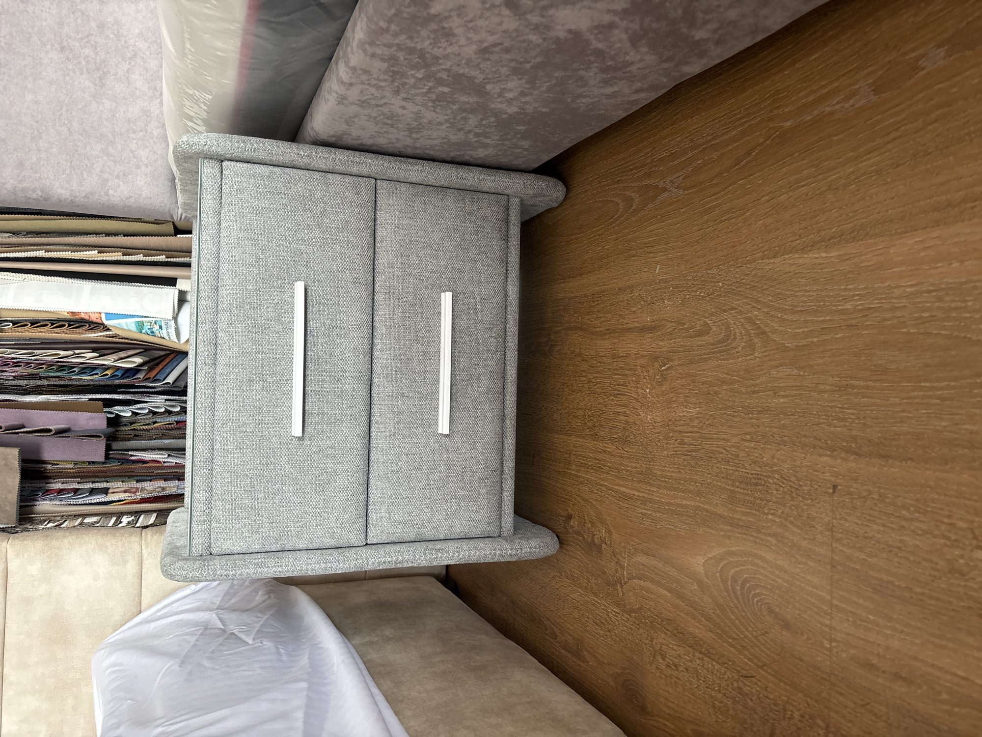 This media file shows a close-up, rotated view of a grey fabric storage cube with two white handles on its front. The cube sits on a light brown wooden floor. To the left of the cube is a stack of fabric swatches, organized in a tiered fashion, with various patterns and colors visible. Behind the fabric swatches, and partially over the storage cube, are large rolls of fabric, covered in plastic wrapping, and a grey textured material. In the foreground, at the bottom of the image, is a beige upholstered surface, partially covered by a white sheet or fabric. The scene appears to be an indoor setting, possibly a fabric store or a storage area for textiles. The lighting is even, suggesting an indoor environment with artificial light, and there are no visible indications of the time of day or weather conditions. No text is visible in the image. The context provided places this scene in Zaporizhzhia, Ukraine.