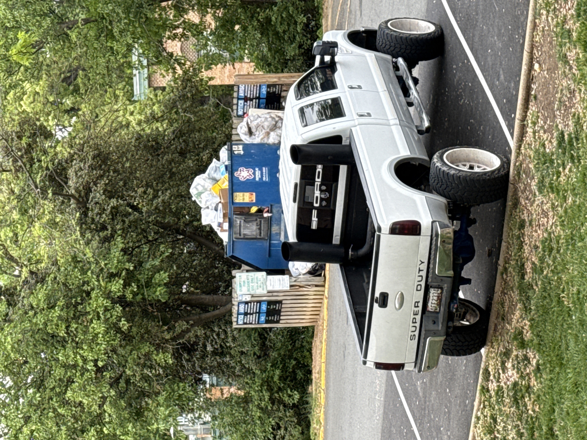 A white Ford Super Duty pickup truck is parked at an angle on an asphalt parking lot next to a grassy patch. The truck has oversized tires and exhaust stacks. A blue dumpster overflowing with garbage is behind the truck. Trees surround the parking area. The license plate is 4FT2134. The location is in Alexandria, United States of America.