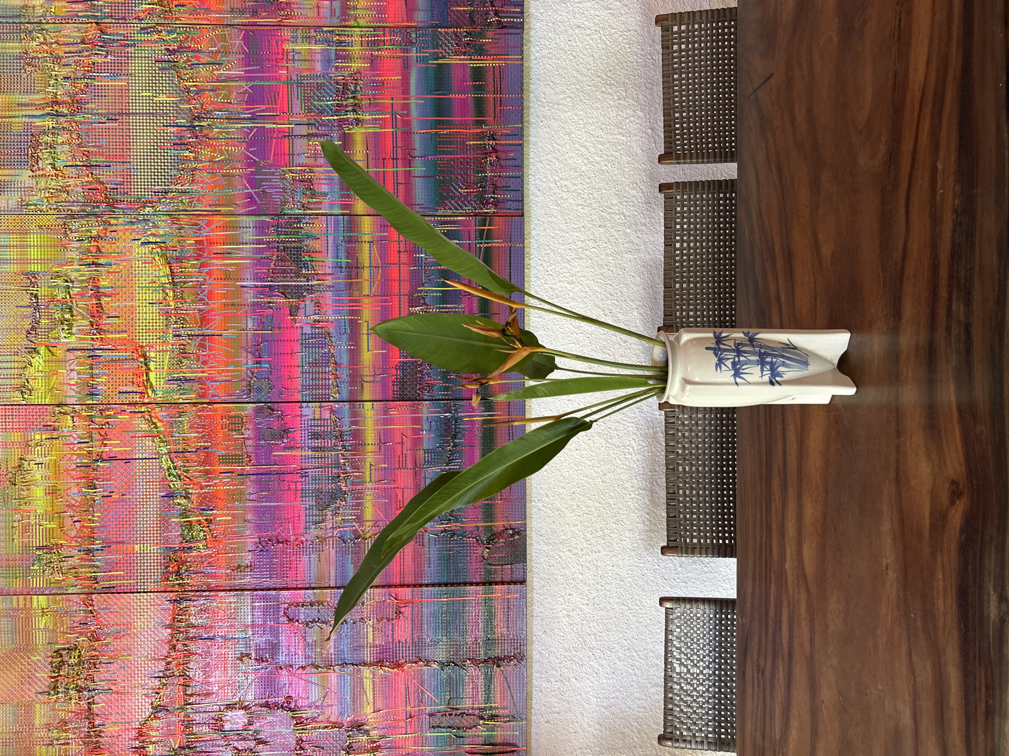 An indoor shot captures a dining table in what appears to be a residence in Mengwi, Indonesia. The table, made of dark wood, dominates the foreground. On it sits a white vase decorated with blue bamboo patterns, containing a bouquet of greenery and orange flowers. Behind the table, three chairs with woven seats and backs are visible. The wall behind the chairs is textured and painted white. A vibrant, abstract artwork composed of multiple panels hangs on the wall, showcasing a kaleidoscope of colors including pinks, yellows, and blues. The overall scene suggests a contemporary and artful interior setting.