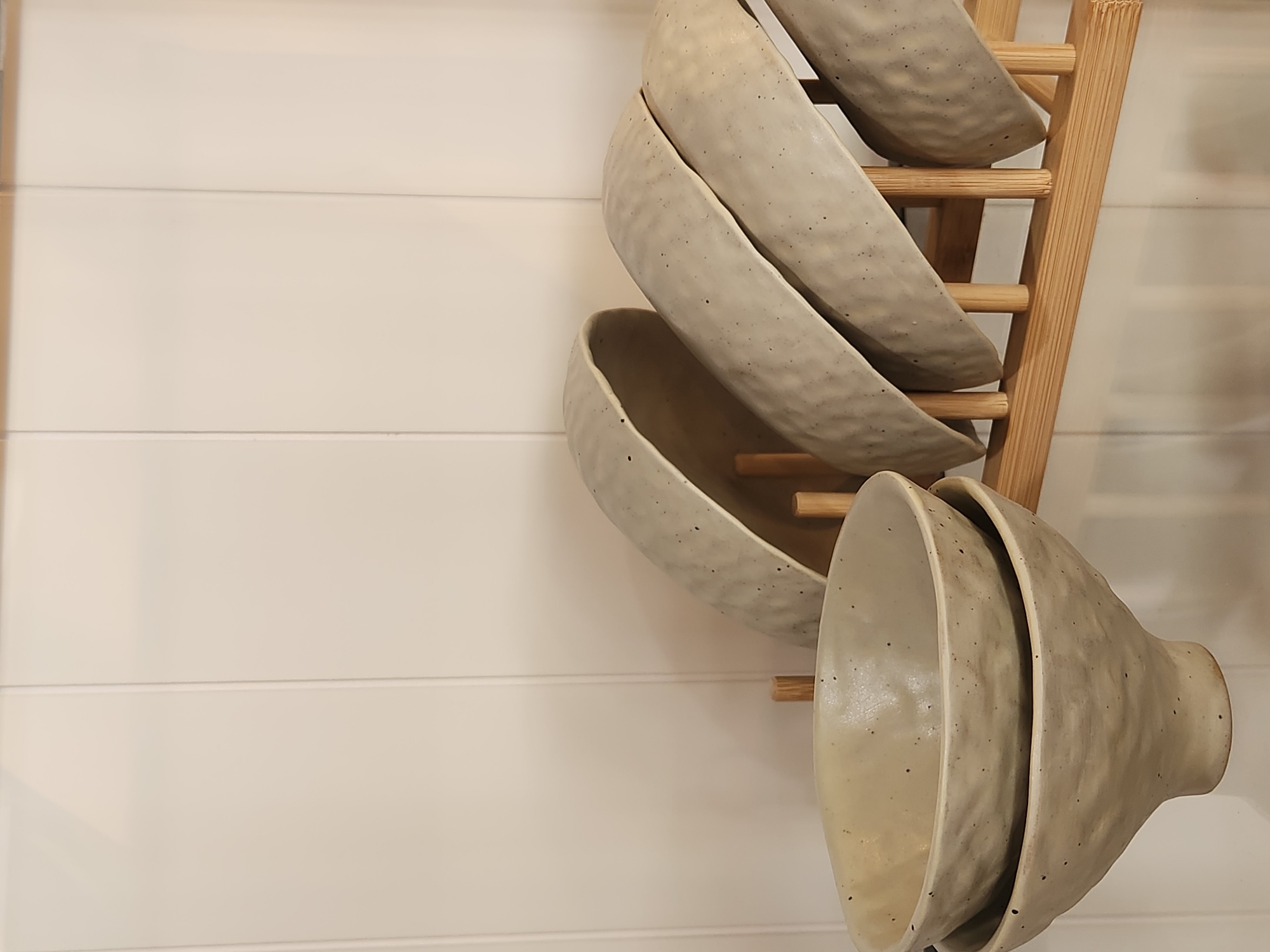 A collection of ceramic bowls are neatly stacked on a wooden rack. The bowls are light in color with a speckled texture, and they appear to be handmade. The rack is made of light-colored wood and is leaning against a wall. The wall is paneled with horizontal wooden planks. The scene suggests a home kitchen or a pottery studio. There are no people visible in the image. The lighting is soft and natural, indicating it could be daytime. The overall mood is calm and organized. There is no visible text in the image.