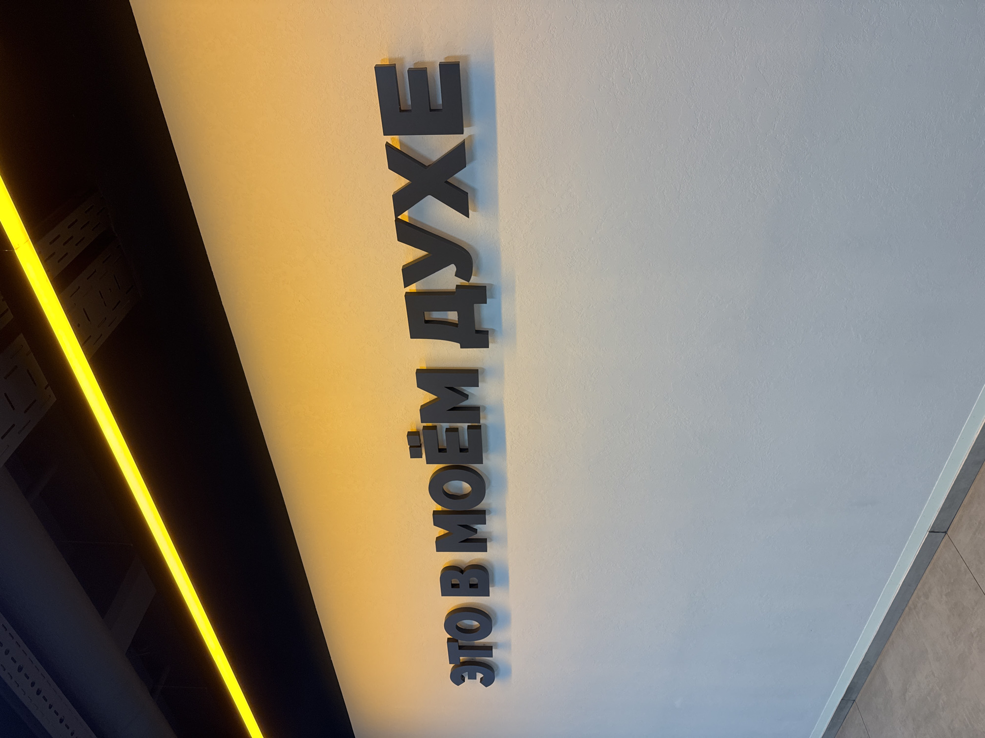 A photograph taken indoors and under artificial lighting features text in Russian on a ceiling. The text reads "ЭТО В МОЁМ ДУХЕ", which translates to "It's in my spirit". The letters are large, dark gray, and possibly made of metal or a similar material. The ceiling is mostly white, with a slight yellow hue from the artificial light source.
To the left, there is a dark structure with a bright yellow linear light fixture running along its edge. This suggests the photo was taken in a modern interior, possibly a commercial or public space in Saint Petersburg, Russia. A partial edge of wall can also be seen to the right. There are no people visible in the photograph.