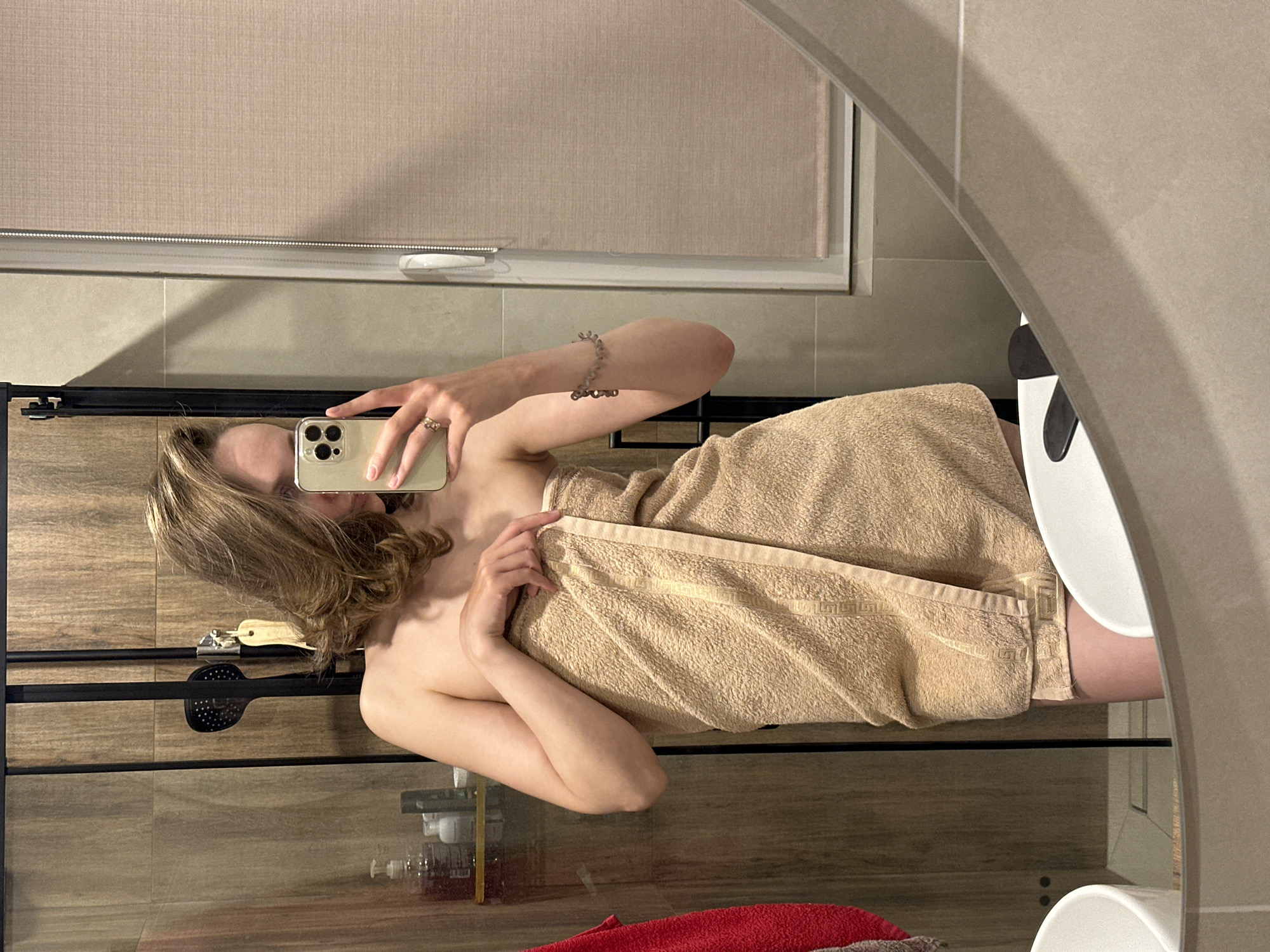 A woman is posing for a mirror selfie in a bathroom. She is holding a smartphone in her right hand, which covers part of her face. She is wrapped in a tan-colored towel. A bracelet is on her right wrist, and a ring is on her right hand. The bathroom has light-colored walls, a window with a closed blind, a black metal towel rack, and a shower with a glass door. The location is a bathroom, likely in a private residence in Sepych, Russia.