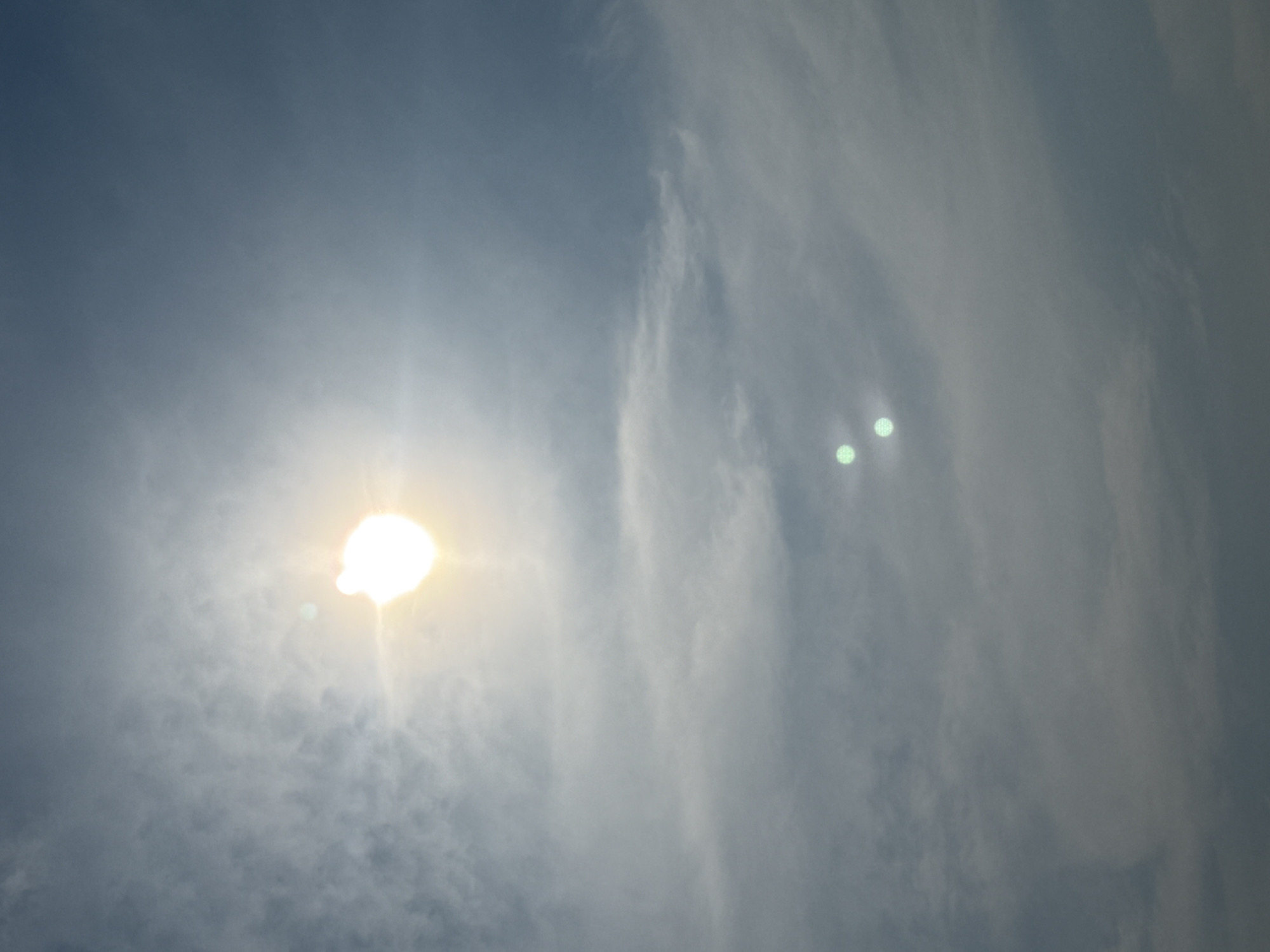 The image depicts a sky with scattered clouds and the sun. The sun is the brightest object, appearing as a white orb with a yellow halo, situated in the upper left quadrant of the image. There are two distinct, circular, greenish-white lens flares in the lower center of the frame. The sky is a gradient of blue and gray, with lighter clouds in the upper portion and denser, grayer clouds lower down. There are no people, animals, or man-made structures visible in the image. The scene is devoid of any specific identifiable landmarks that would indicate the city of Norilsk, Russia.