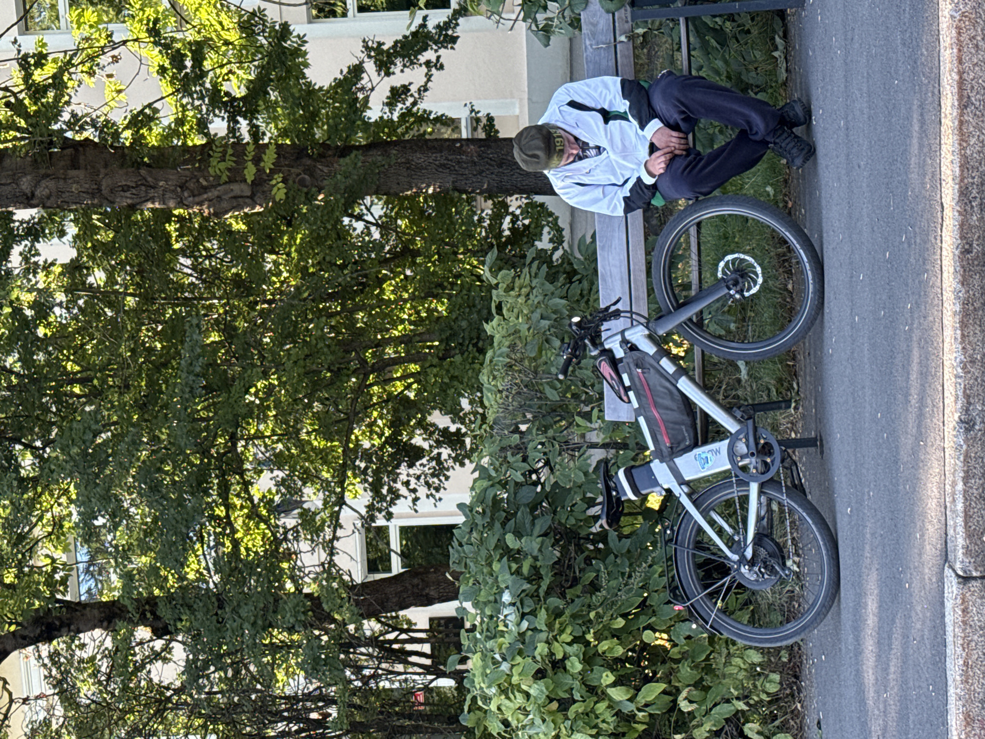 A person wearing a green beanie and a white and green jacket is sitting on a wooden bench, with their head bowed and hands clasped. Their dark pants and black shoes are visible. An electric bicycle, silver with black accents and a red bag attached, is parked to the left of the bench. The scene is outdoors, with lush green bushes and trees providing a backdrop. Sunlight filters through the leaves, creating dappled light. In the background, a building with light-colored walls and windows is visible. The ground in front of the bench is paved with asphalt. This image was captured in Tallinn, Estonia, likely in a park or a public area with seating and greenery.