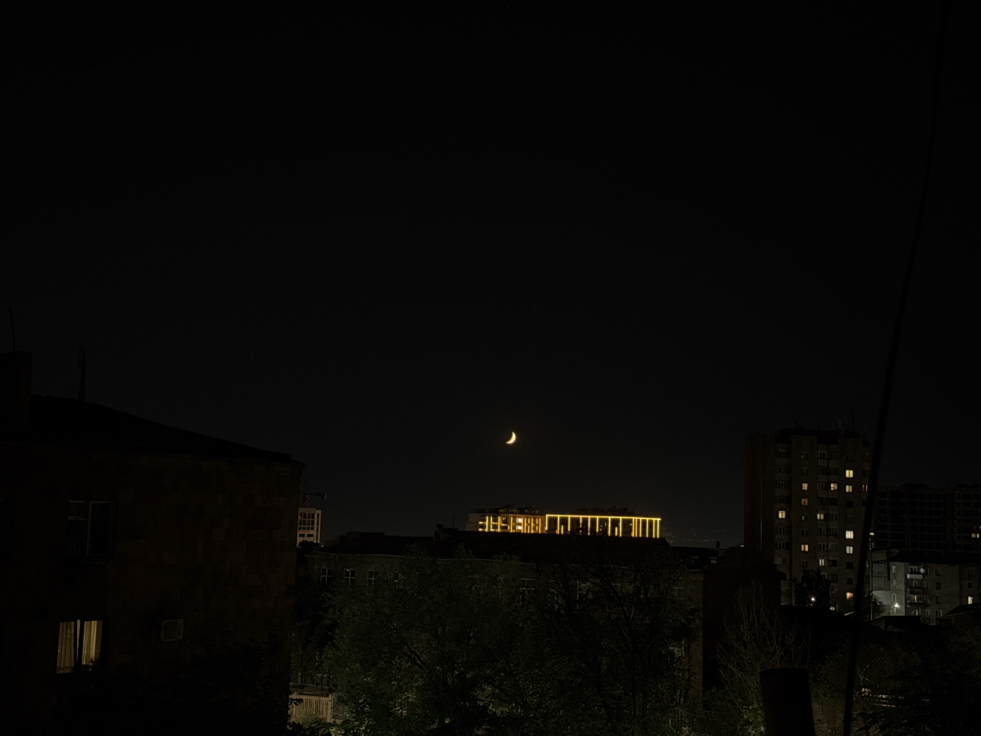 The image captures a nocturnal cityscape in Yerevan, Armenia. In the foreground, a dark, textured building with lit windows occupies the left side. To the right, trees partially obscure lower buildings. In the midground, a low-rise building is visible with a distinctive yellow, illuminated architectural feature across its roof. Further back, a tall residential building stands out with numerous lit windows, suggesting occupancy. The sky is dark, featuring a visible crescent moon. The scene conveys a typical urban night environment.