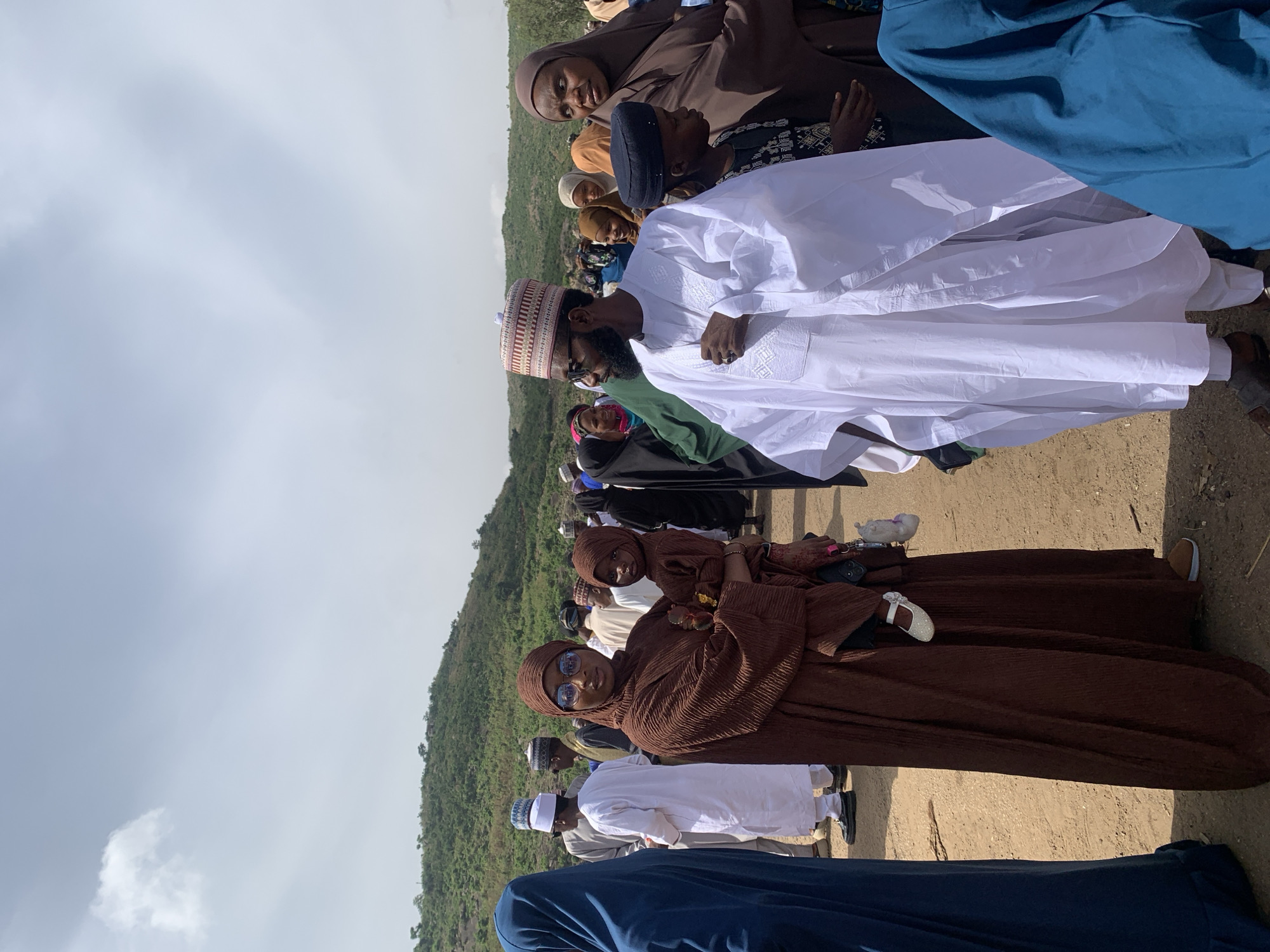 People stand outdoors on a dirt surface near a hill. The sky is cloudy. Many people wear traditional Muslim clothing and head coverings. Some wear glasses. One person holds a child. Naraguta Hausawa, Nigeria is not visible.