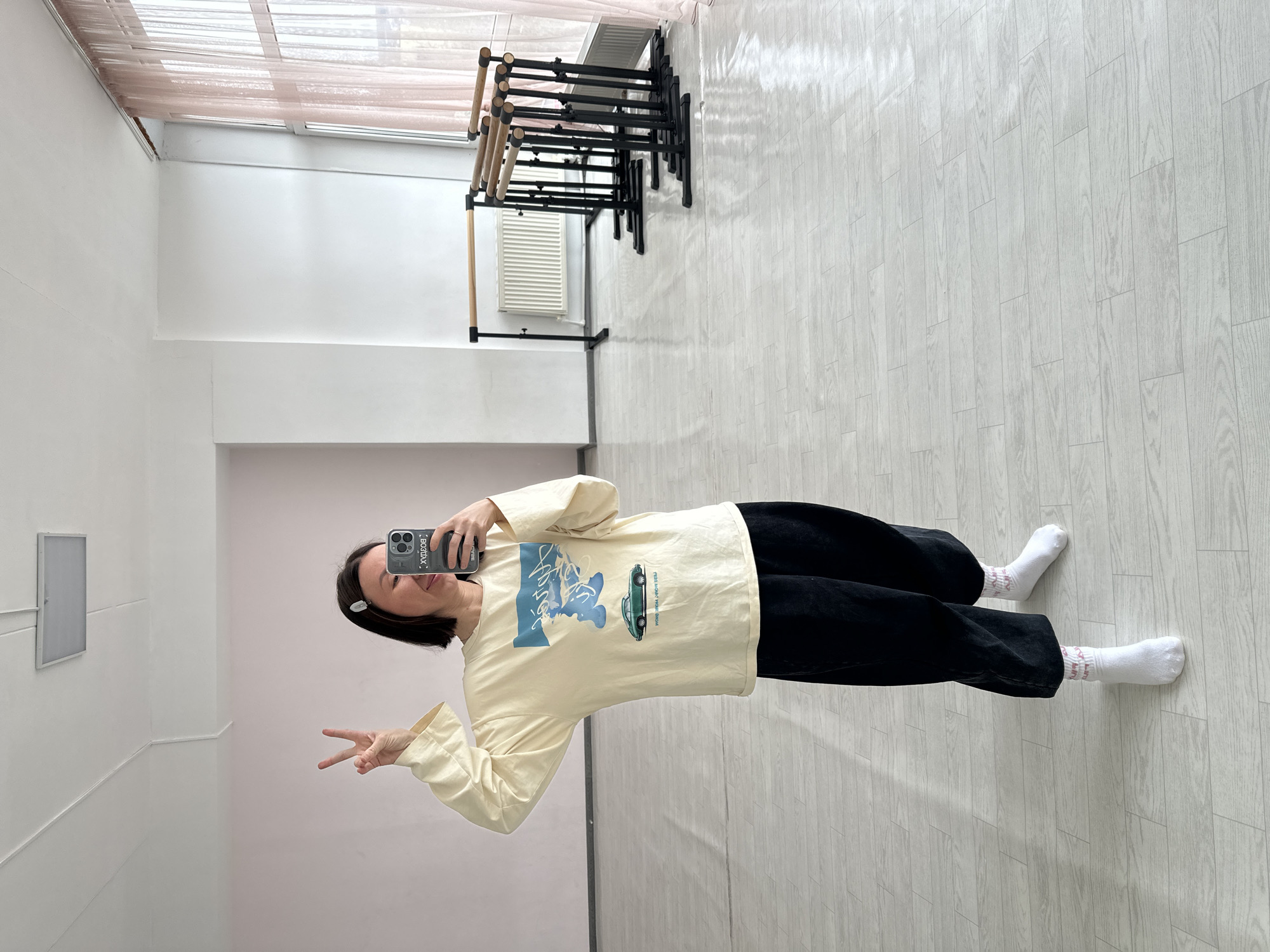 A woman takes a mirror selfie in a well-lit indoor space, likely a dance studio, in Votkinsk, Russia. She is positioned centrally in the frame, holding a smartphone in her right hand to capture her reflection while making a peace sign with her left hand. She has dark, chin-length hair with a white clip on her right side and a subtle smile.
She is dressed in a light yellow long-sleeve shirt featuring a blue graphic design that includes stylized text and a green vintage-style car; a portion of the text reads "must-have Your Honda." She wears black pants and white ankle socks with faint pink writing. Her black phone case is visible, displaying the white text "BODBAX.".
The background features white walls and a light-colored wooden floor with a plank pattern. On the right side of the room, several black-framed ballet barres with wooden rails are stacked or arranged against the wall. Above them, a long window with translucent, light-colored blinds allows natural light into the room. A small, square, light-colored fixture, possibly a vent or access panel, is mounted on the left wall. The bright lighting suggests it is daytime.