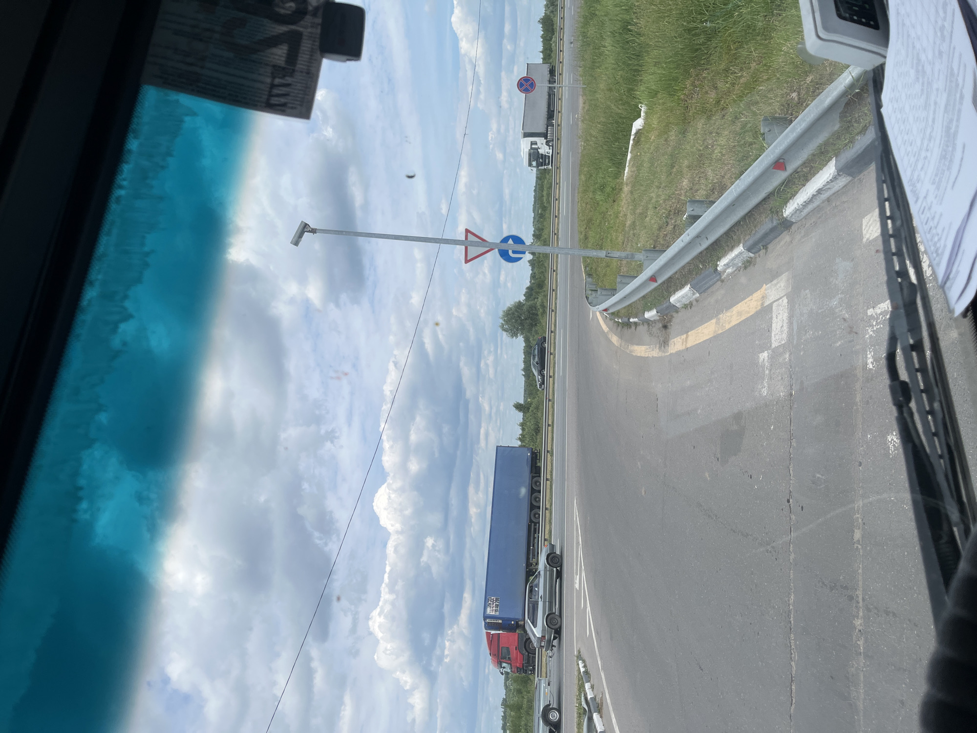 The image is taken from inside a vehicle, looking forward through the windshield. The scene is a road intersection in Старое Бобренево, Russia, under a cloudy sky. On the left, a grey passenger car is seen driving on a paved road. In the middle ground, a large red truck with a blue trailer is driving on a highway. Further to the right, another white truck with a grey trailer is also on the highway. Road signs are visible, including a give way sign, a blue directional arrow sign, and a red and white circular prohibition sign. Guardrails line the side of the road, and tall green grass and trees are visible in the background. The interior of the vehicle shows a dashboard with papers and a piece of equipment.