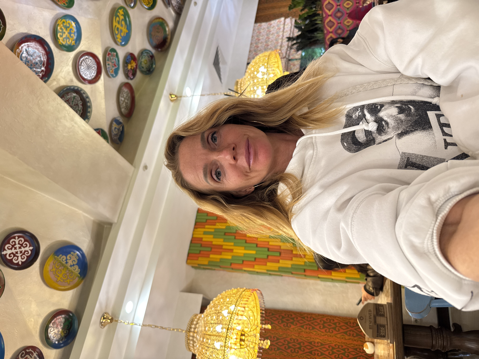 An indoor, vertical selfie shows a blonde woman with blue-grey eyes looking directly at the camera with a neutral expression. She is wearing a white hooded sweatshirt featuring a black and white graphic of a person's face, possibly male, with dark hair, and a partial letter "E" visible next to it. She has small, subtle hoop earrings.
The background is richly decorated, suggesting a restaurant or upscale establishment in Bishkek City, Kyrgyzstan. On the left, a light-colored wall is adorned with numerous circular decorative plates of various sizes, colors (blue, yellow, red, green, white, black), and intricate, often symmetrical, patterns. One plate clearly displays a blue and yellow design with a yellow motif.
In the mid-ground and background, ornate chandeliers with multiple bulbs and glass or crystal elements hang from the ceiling, casting a warm glow. A section of wall or a partition behind the woman features a colorful, geometric pattern composed of stacked rectangular elements in shades of green, orange, yellow, and red. Below this, a patterned textile or rug with red and orange geometric designs is visible.
To the right, a dark wooden table is partially seen with a "Reserved" sign clearly visible on it. A small, white object rests next to the sign. Part of another person's arm/hand is also faintly visible in the background, near the table. The lighting suggests an indoor setting, possibly evening or a well-lit daytime interior.