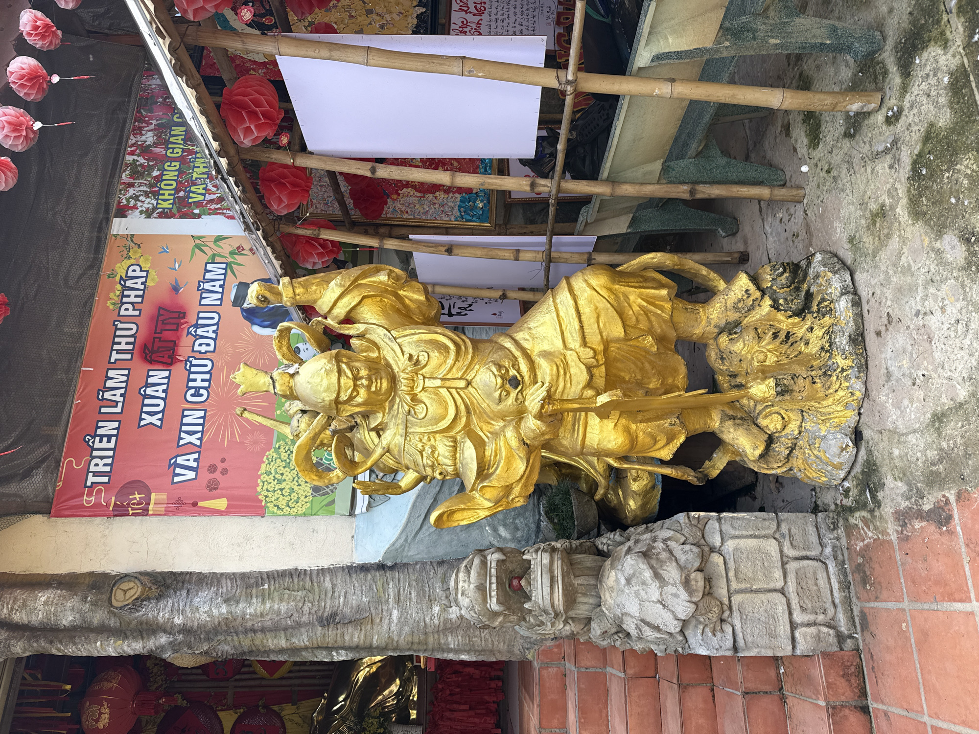 The image depicts an outdoor scene in Lâm Viên - Đà Lạt, Vietnam. The central focus is a large, golden statue of a warrior figure, adorned with armor and a headdress, holding a sword. The statue stands on a rocky base. To the left of the statue, there is a stone pillar topped with a dragon-like carving, and a red-tiled staircase leading upwards. Behind the statue, a large red banner with Vietnamese text and images of birds and flowers is visible. Further back, a structure with a bamboo roof and several red paper lanterns are present. The ground is paved with terracotta tiles in some areas and concrete in others, with some patches of greenery and debris. The overall environment appears to be a temple or shrine.