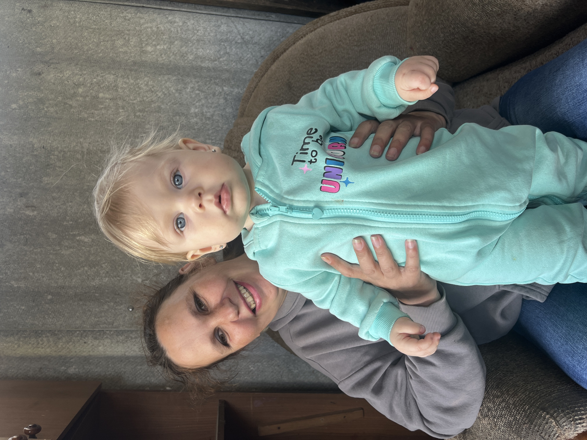 A woman sits on a brown upholstered chair, holding a baby. The woman is wearing a gray sweatshirt and blue jeans. The baby is wearing a light blue zippered onesie with the words "Time to be Unicorn" on it. The background is a gray wall. It is impossible to identify the location as Фоки, Russia.