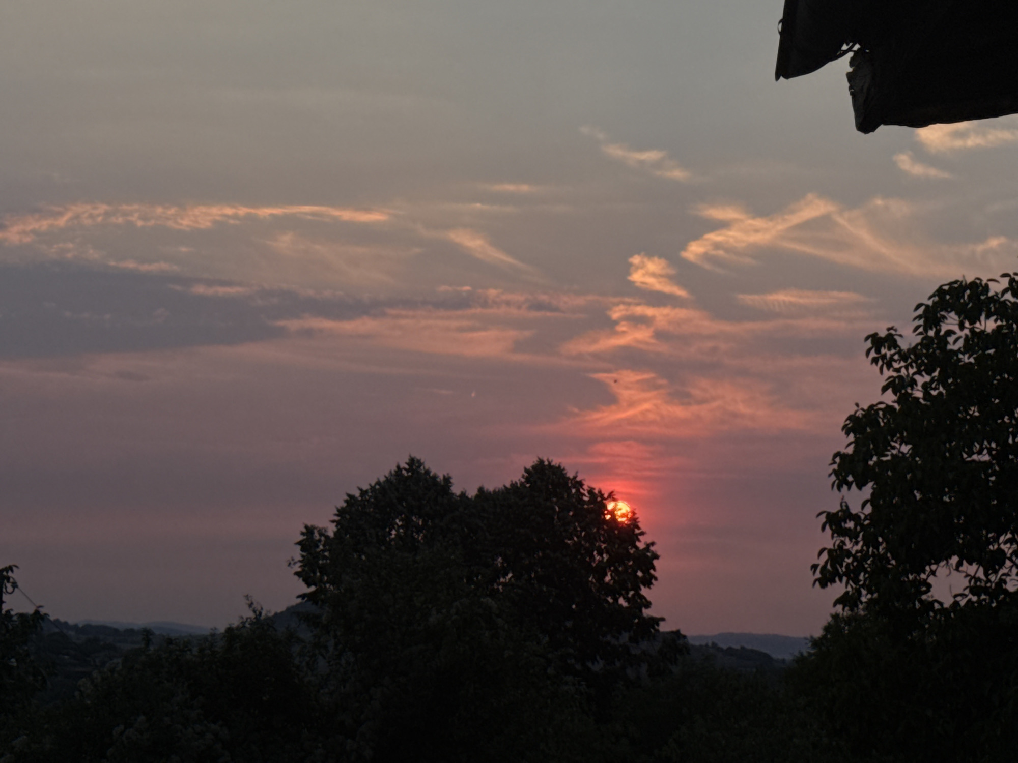 This is a landscape photograph taken at sunset in Krushevo, Bulgaria. The sun is partially obscured by a large tree in the foreground, creating a vibrant red glow. The sky is filled with streaks of pink and orange clouds. In the distance, rolling hills are silhouetted against the colorful sky. A dark, overhang structure is visible in the top right corner of the image, possibly part of a building or shelter. The overall scene depicts a serene and picturesque sunset.