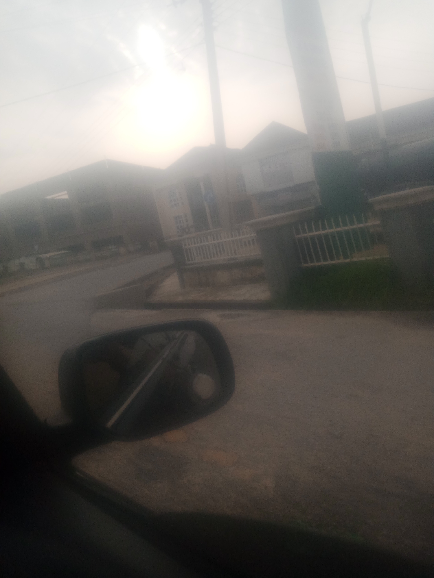 The image depicts a street scene in Jalingo, Nigeria, characterized by overcast skies and low visibility, possibly due to fog or haze. In the foreground, a portion of a car's interior is visible, including the side mirror reflecting a blurry image of the car's interior and the surrounding environment. The street itself appears to be paved, with a visible curb and drainage system. Buildings line the street, some appearing to be residential or commercial structures with multiple stories. A white metal fence with vertical bars is situated along the side of the road, bordering a patch of green grass. Utility poles and overhead wires are also present, indicating the presence of infrastructure. The overall impression is of a typical urban environment under subdued lighting conditions.