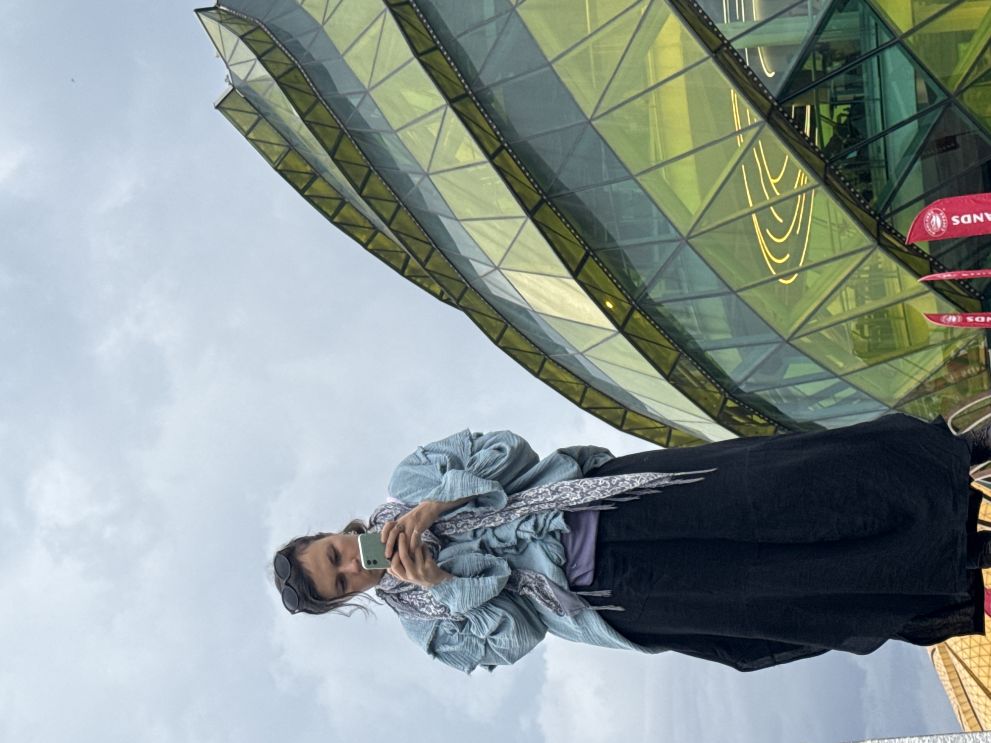 A woman stands looking at her smartphone against the backdrop of a modern architectural structure with glass and geometric designs. The sky overhead is overcast. The woman wears a long black garment and a blue shirt, and her sunglasses are on her head. The building behind her has a green and yellow glass facade. In the upper right corner, parts of a flag with "Sands" printed on it are visible. Based on architectural design and flags, the photo appears to be in front of the Grand Lisboa Palace in Da Lat, Vietnam.