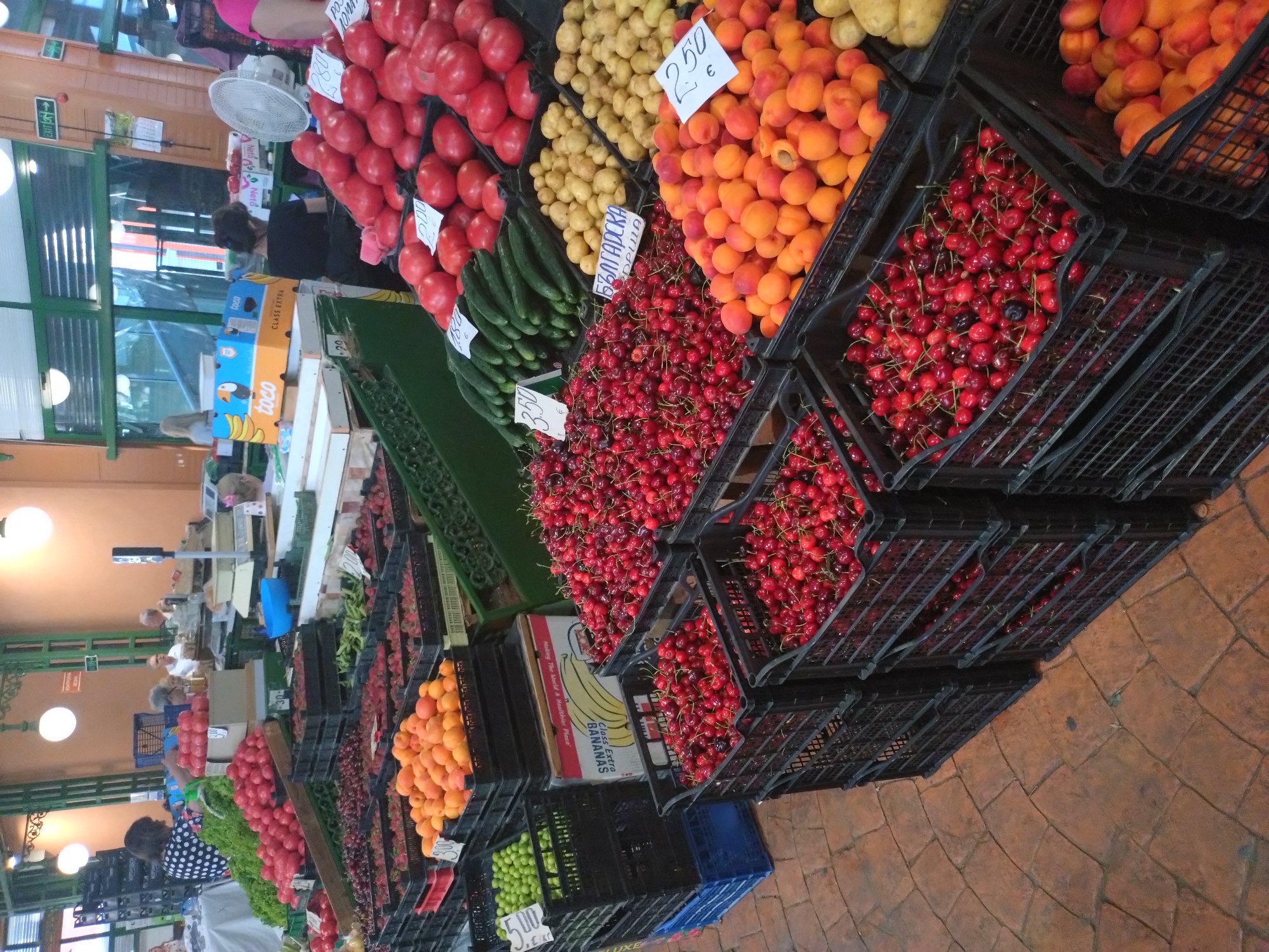 The image depicts a vibrant indoor produce market in Stara Zagora, Bulgaria. Various fruits and vegetables are displayed in black plastic crates, including tomatoes, cucumbers, potatoes, apricots, cherries, oranges, green apples, and bananas. Price tags are visible near the produce. People are present both behind and in front of the stalls. The flooring is made of reddish-brown stone-like blocks. A fan is visible on one of the stalls. Boxes of bananas are stacked among the crates. There is also what appears to be an exit sign at the top left of the image.