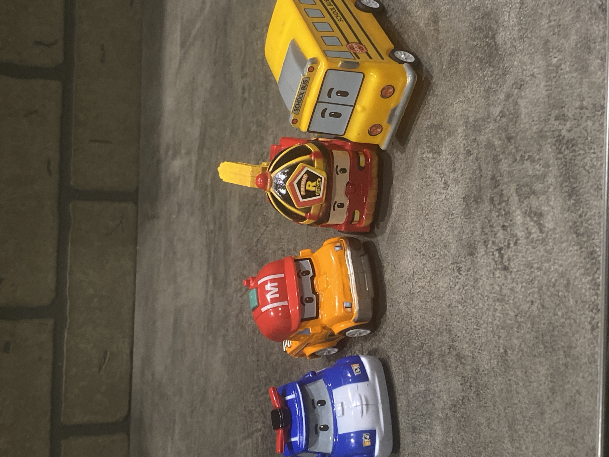 The image shows four toy cars lined up on a gray, textured surface. From top to bottom, the cars are a yellow school bus, a red and black fire truck, an orange tow truck with a red cab, and a blue and white airplane car. The background includes a partial view of a brick wall on the left. The scene does not depict a location in the city of Sochi, Russia.