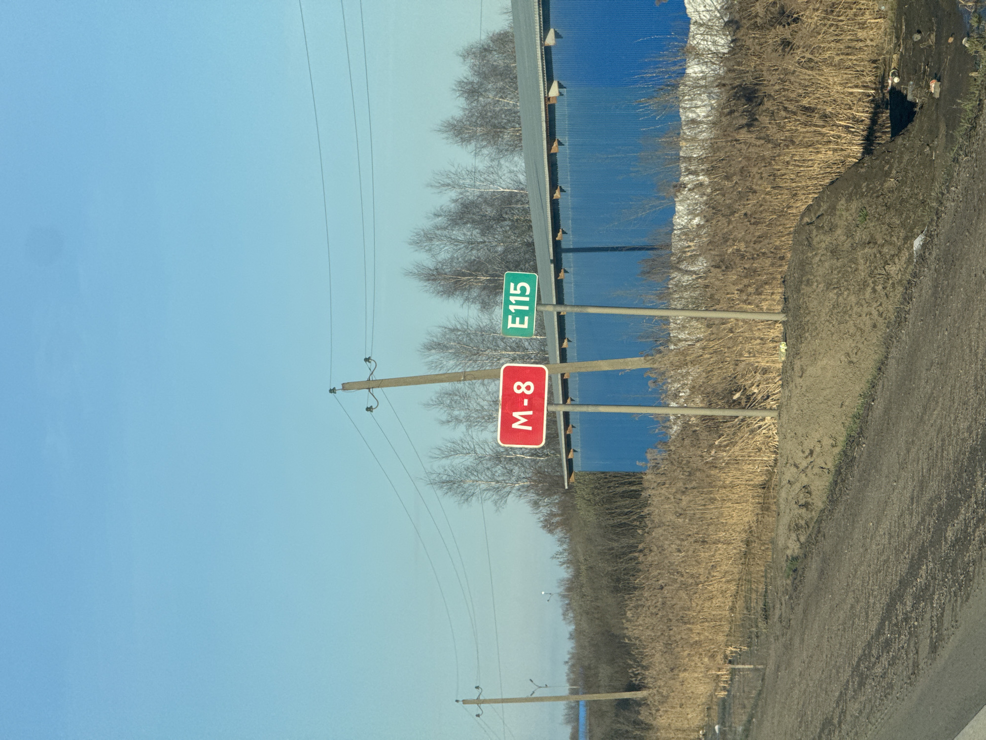 The image depicts a roadside scene in Ново-Никольское, Russia. Two road signs are visible: one red with "M-8" in white, and a green one with "E115" in white. These signs are mounted on poles next to a road. A blue corrugated metal wall or barrier runs parallel to the road and the signs, likely serving as a sound barrier or boundary marker. To the right of the blue wall is a dense strip of dry, brownish vegetation. Overhead, utility poles and power lines crisscross the blue sky. The road itself is visible in the foreground, showing a mix of dirt and paved surfaces.