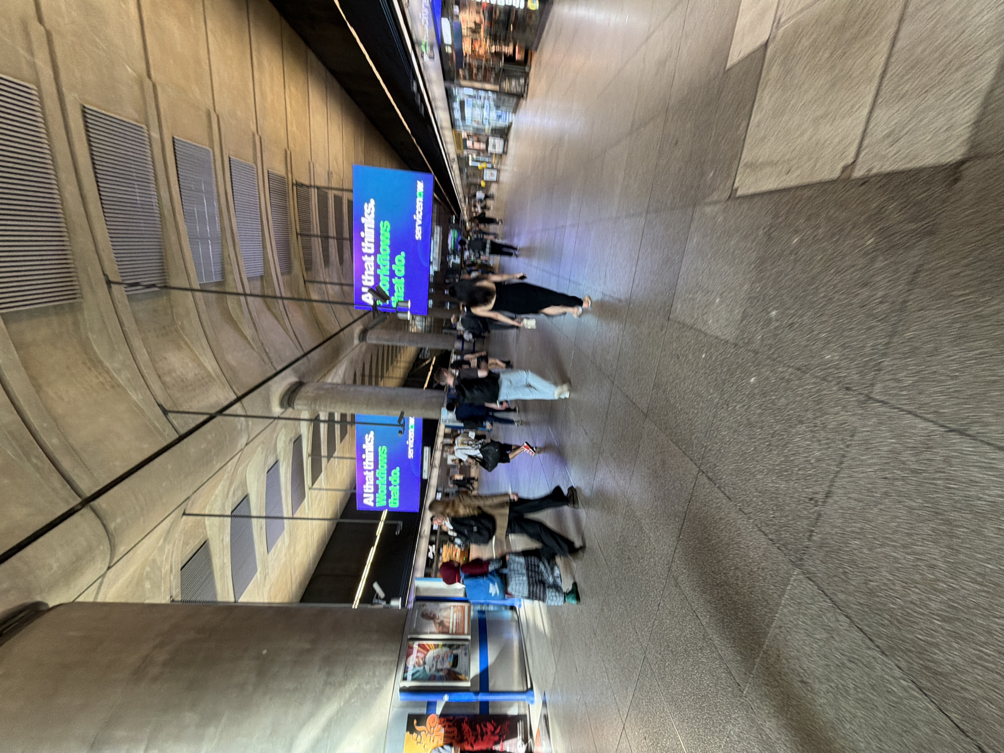 The image captures an interior scene in the city of London, United Kingdom, specifically the Elizabeth Line platform at Tottenham Court Road Station. Several people are visible, walking along the platform. There are two illuminated advertisements for ServiceNow with text "Al that thinks. Workflows that do." The architectural structure of the station is emphasized by the curved walls and repetitive vent designs. In the foreground, there is a display case of what appears to be maps or flyers. The overall environment is brightly lit.